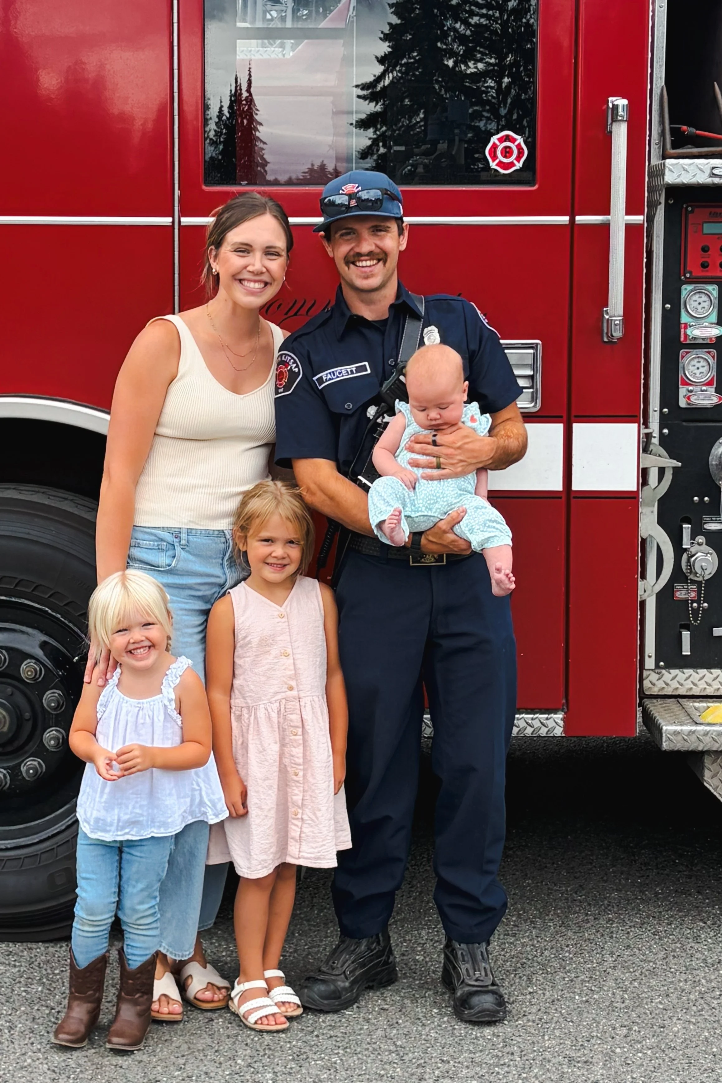 A firefighter in uniform holding a small baby with three young girls smiling in front of a red fire truck.