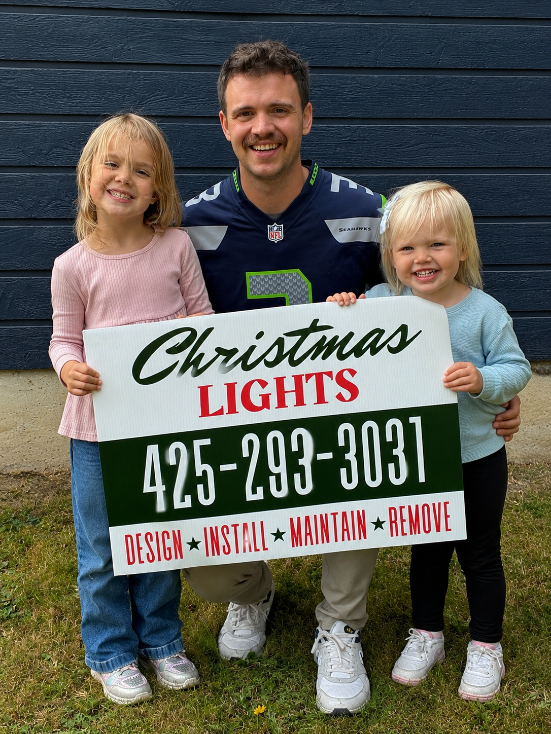 A man in a Seattle Seahawks football jersey standing outside with two young girls, holding a sign that reads "Christmas Lights" along with a phone number and the words "Design, Install, Maintain, Remove." The girls are smiling and wearing casual clothes, standing on grass in front of a dark blue wooden wall.