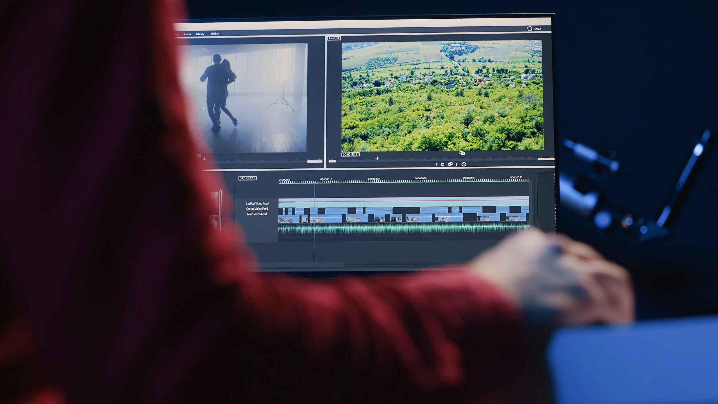 Close-up of a video editing monitor showing video clips and landscape footage, with a person's hand and part of their red clothing visible in the foreground.