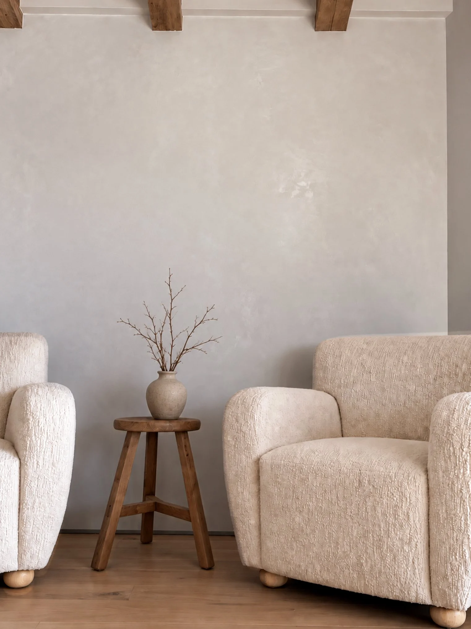 Two cream textured armchairs beside a natural wood stool and ceramic vase against a soft plaster wall with exposed ceiling beams.