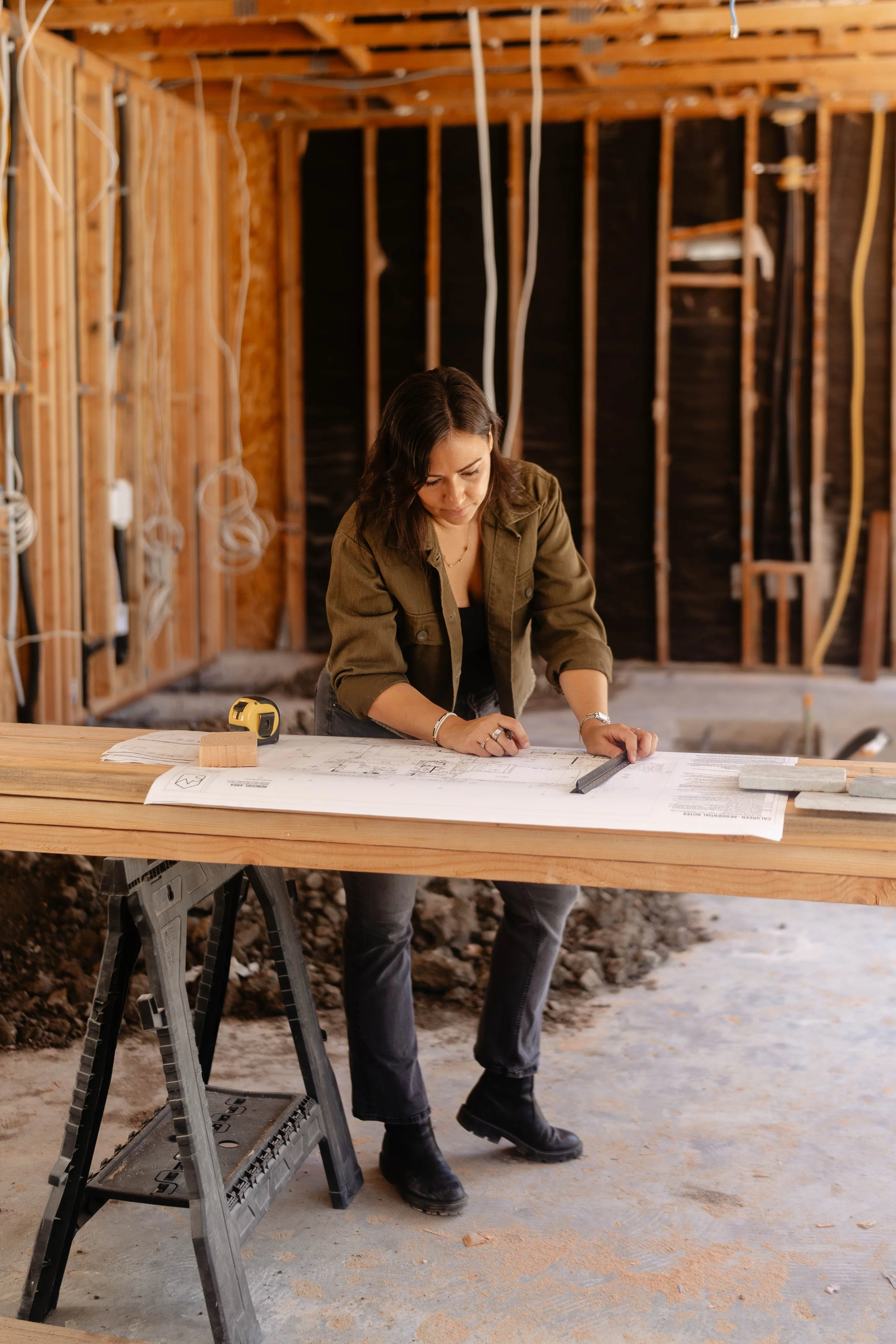 Woman working on blueprints at a construction site, with exposed wall framing and construction tools on a work table.