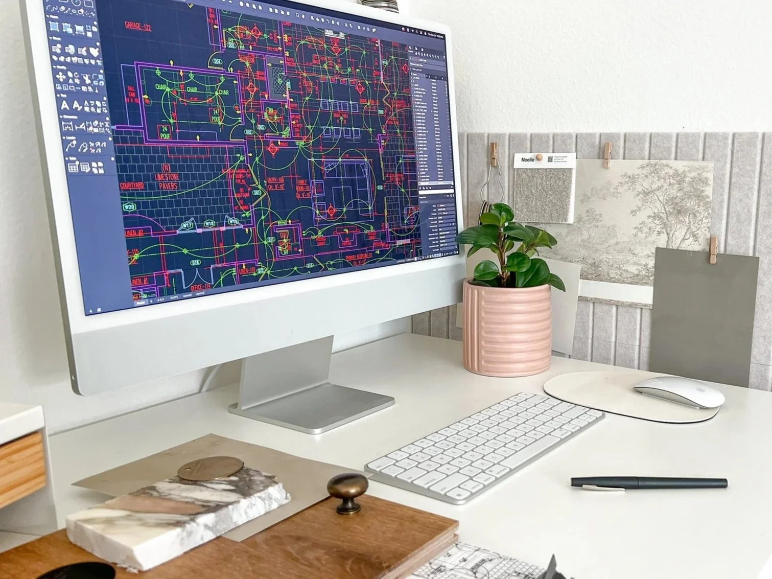 Desktop computer display showing a detailed architectural or engineering blueprint with colorful lines and symbols, on a white desk with a white keyboard, mouse, a pink plant pot with a green plant, and some framed artwork pinned to a wall.