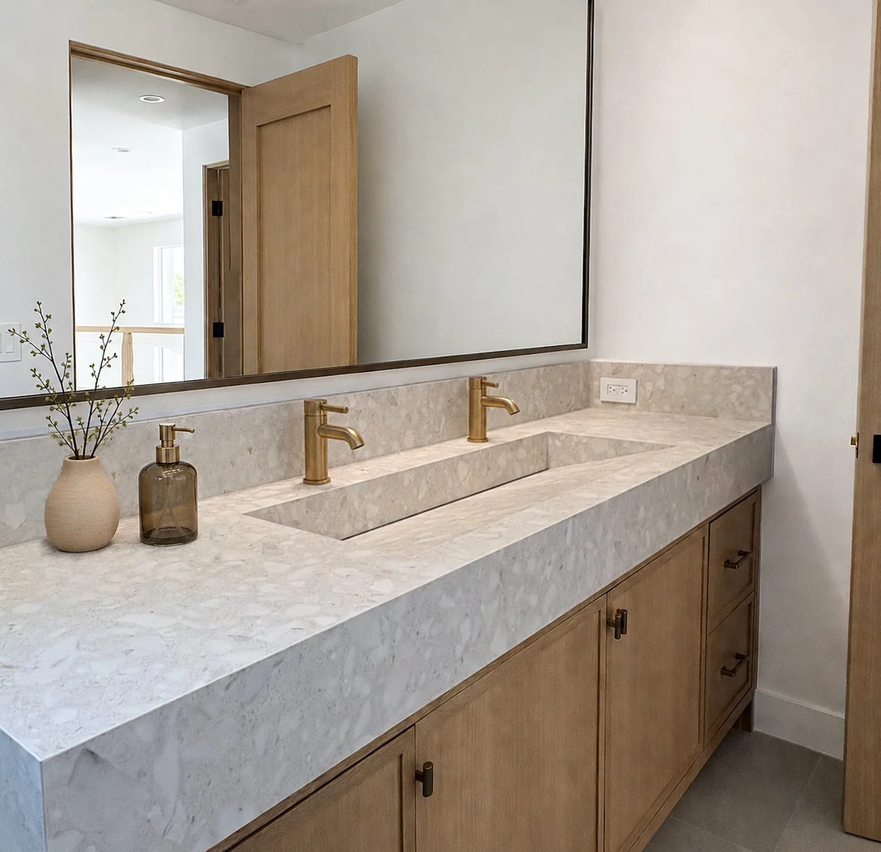 Bathroom vanity with beige marble countertop, two gold faucets, a large mirror, a small vase with a branch, a soap dispenser, and wooden cabinetry.