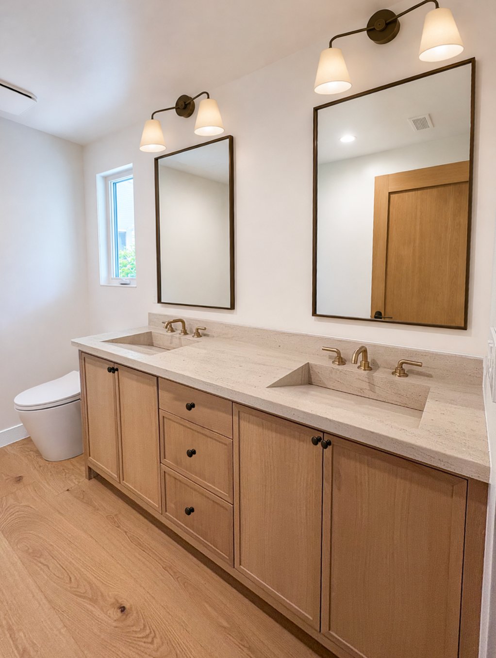 Bathroom with double vanity, two mirrors, wall-mounted lights, wooden cabinetry, beige countertop, and toilet, with a small window.