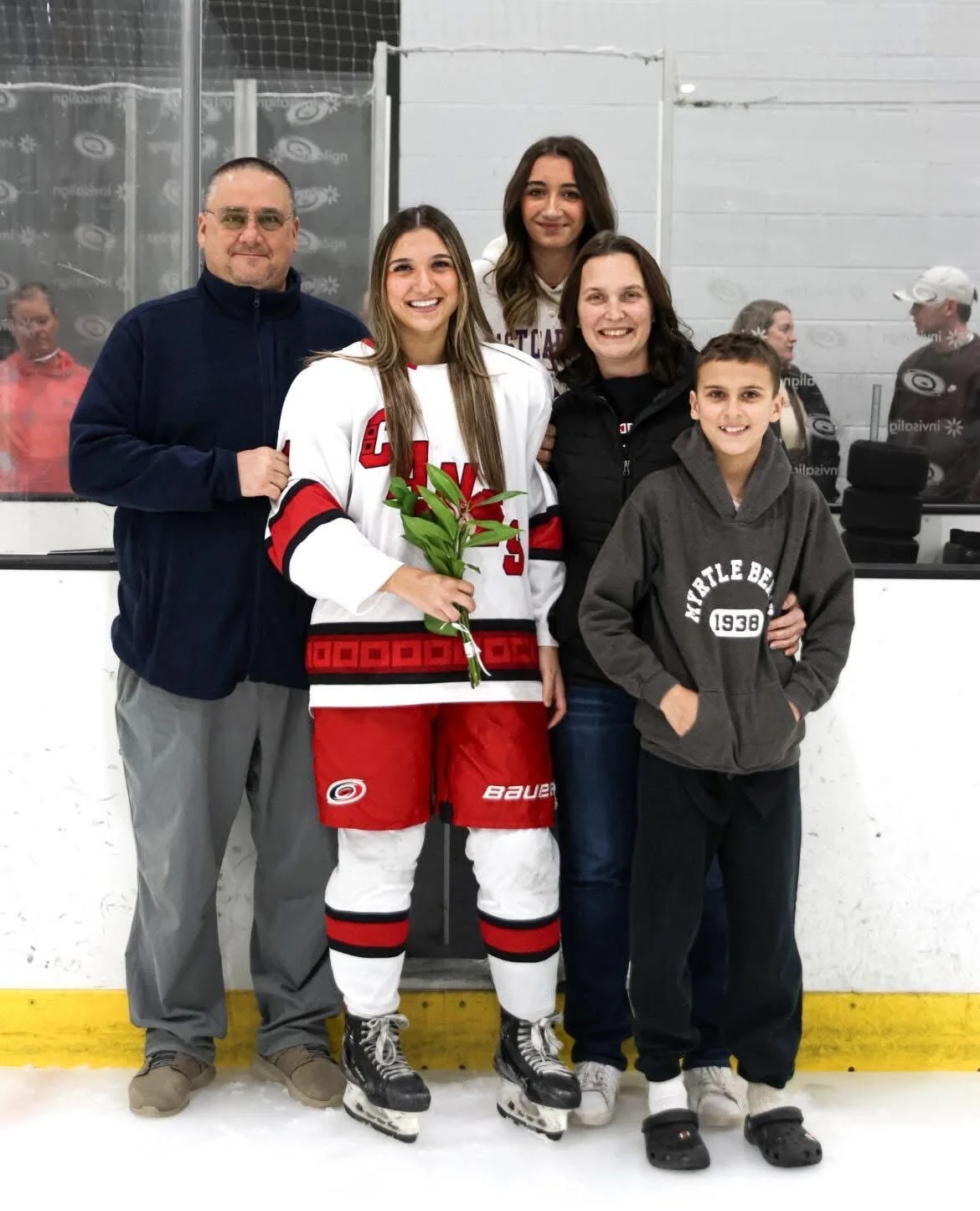 A girl in hockey gear holding a bouquet of flowers, surrounded by family members at an ice rink.