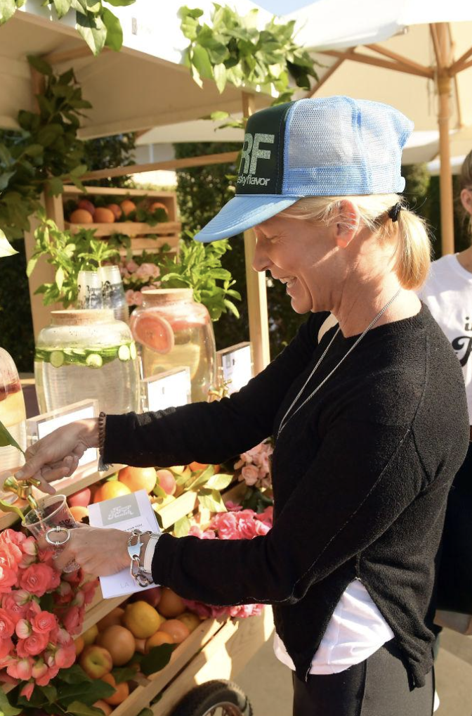 Woman smiling and shopping for fruits and flowers at an outdoor market stand, wearing a blue cap and a black sweater.