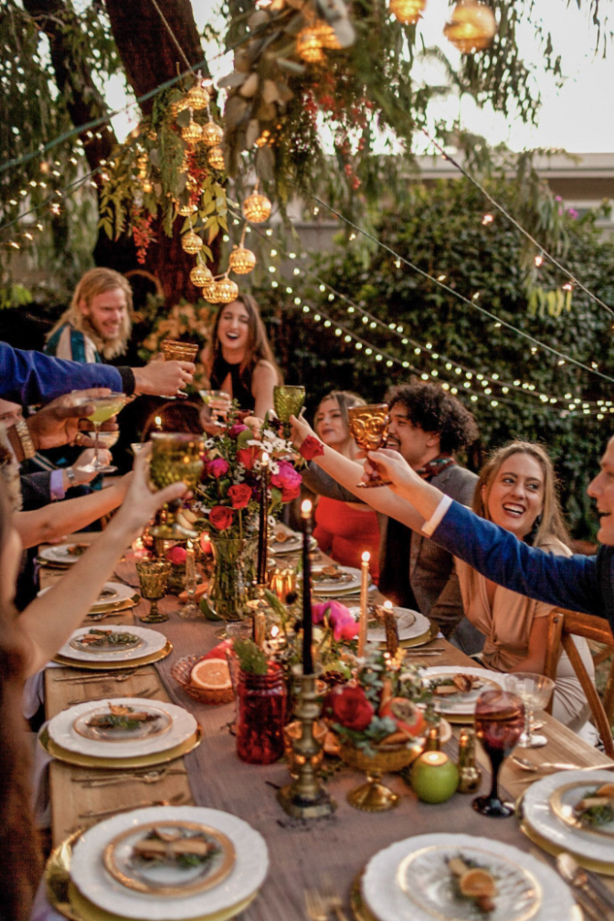 People celebrating at a festive dinner party outdoors, raising glasses in a toast, with decorated table, flowers, candles, and hanging lights.