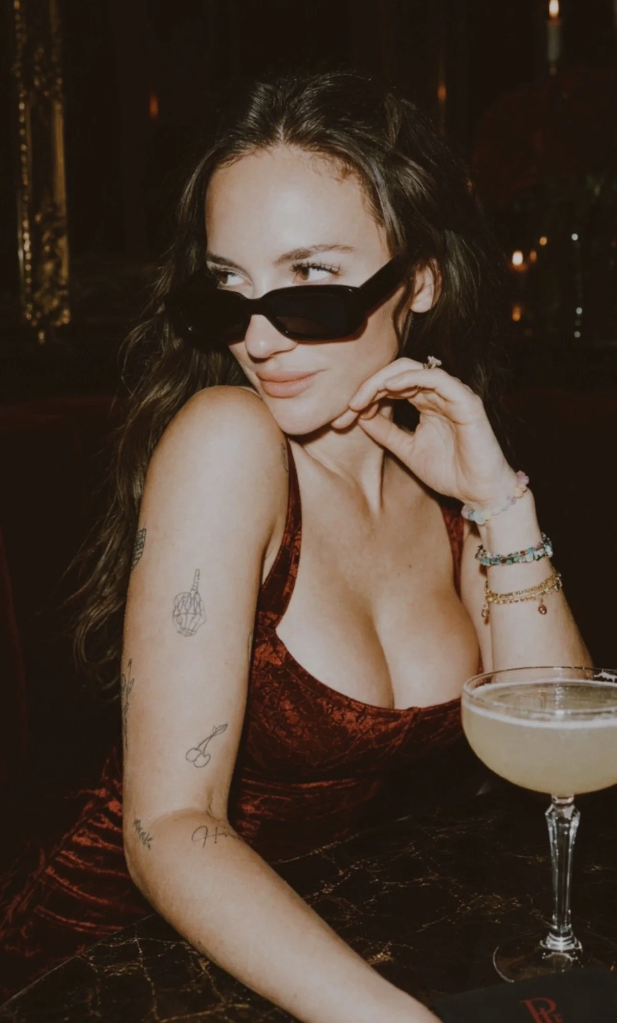A young woman with long dark hair wearing sunglasses, a red patterned dress, and multiple bracelets sitting at a bar or restaurant. She has tattoos on her left arm and is smiling slightly, holding her chin with her left hand. There is a cocktail glass with a white drink in front of her.