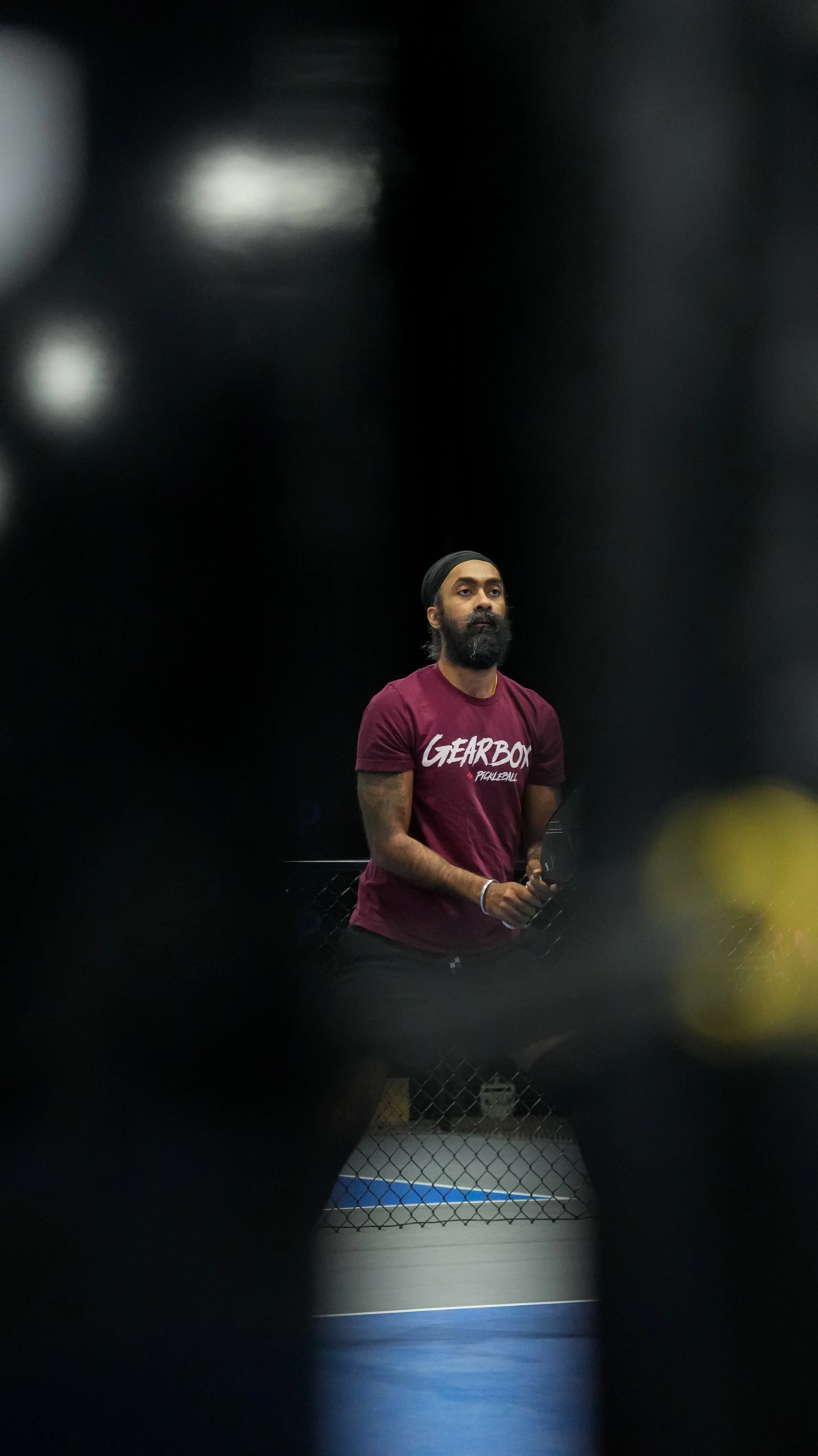 A man with a beard and black head covering standing inside a cage with chain-link fencing, wearing a maroon t-shirt that says 'GEARBOX PICKLEBALL'.
