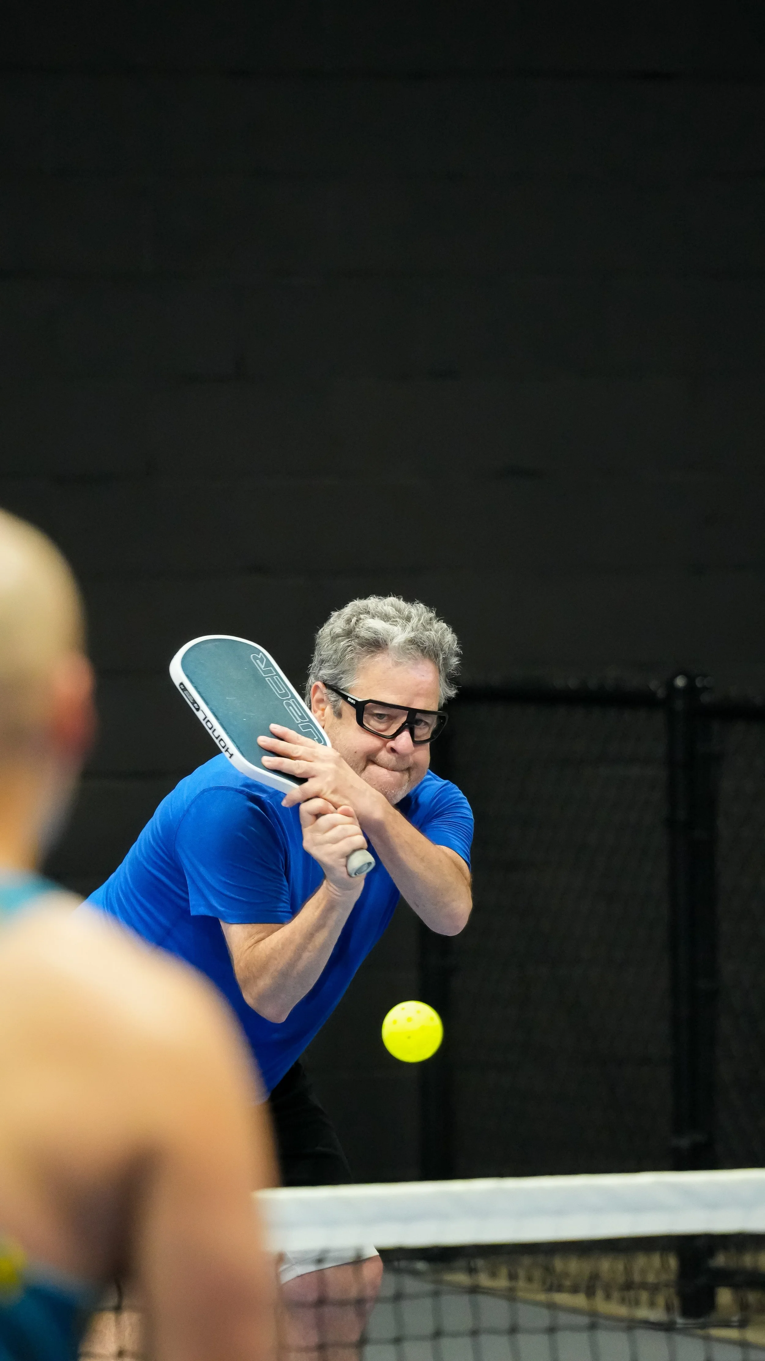 A middle-aged man wearing glasses and a blue shirt hitting a yellow tennis ball with a racket during a game, indoor tennis court with black walls.
