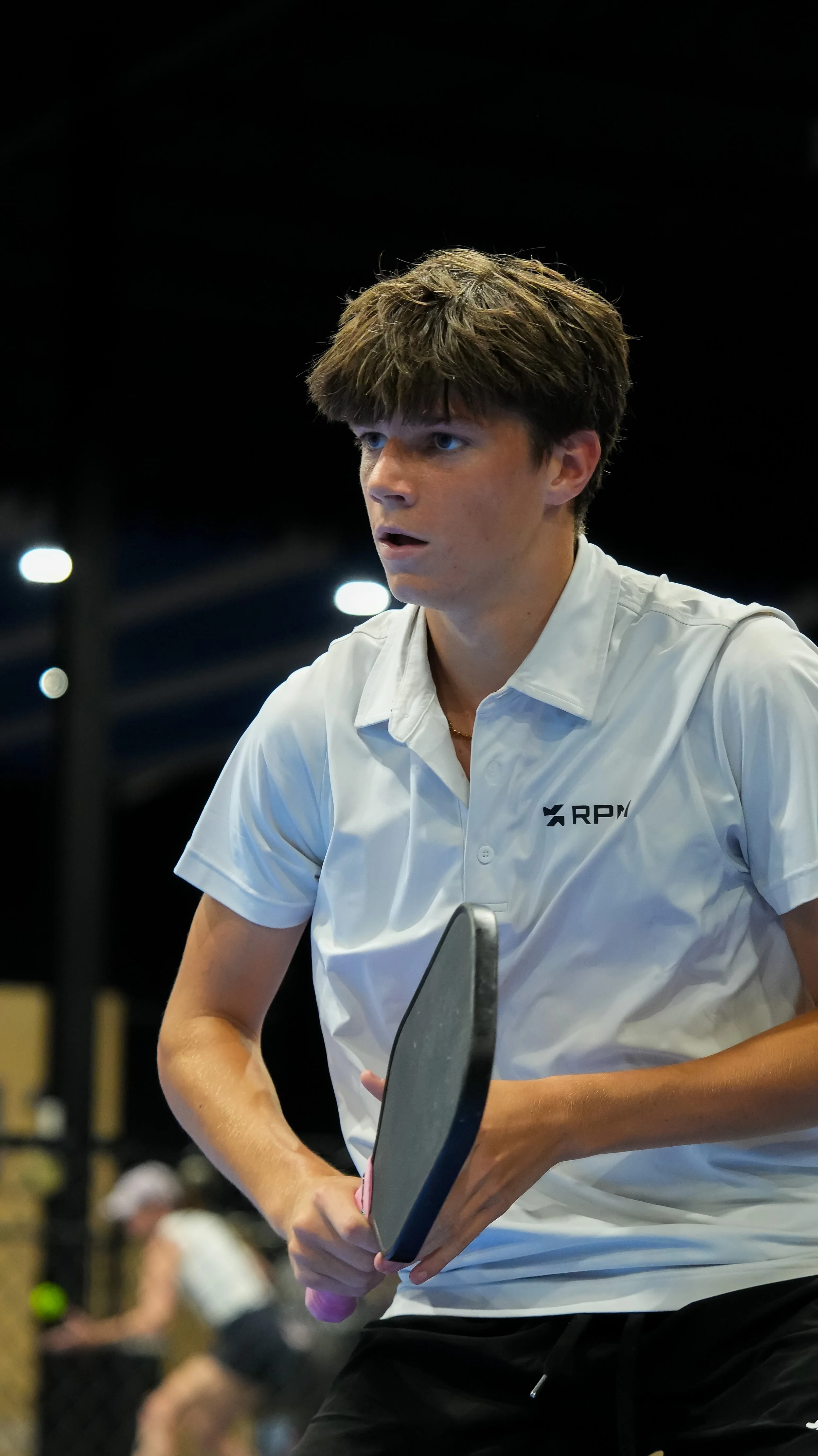 Young man with brown hair wearing a white shirt holding a ping pong paddle in an indoor setting with other people in the background.