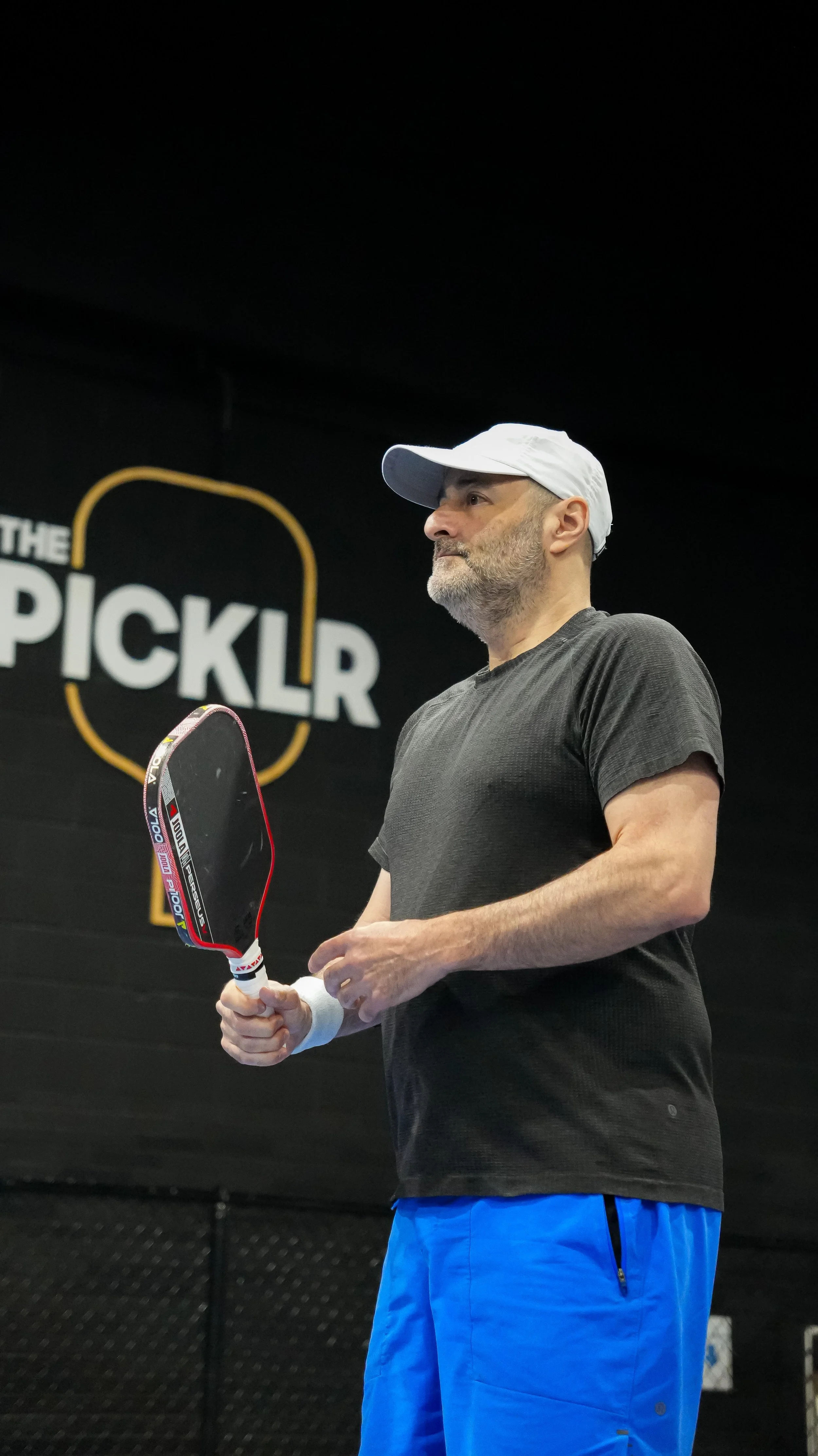 A man wearing a white cap, black t-shirt, and blue shorts holding a pickleball paddle at an indoor pickleball court.