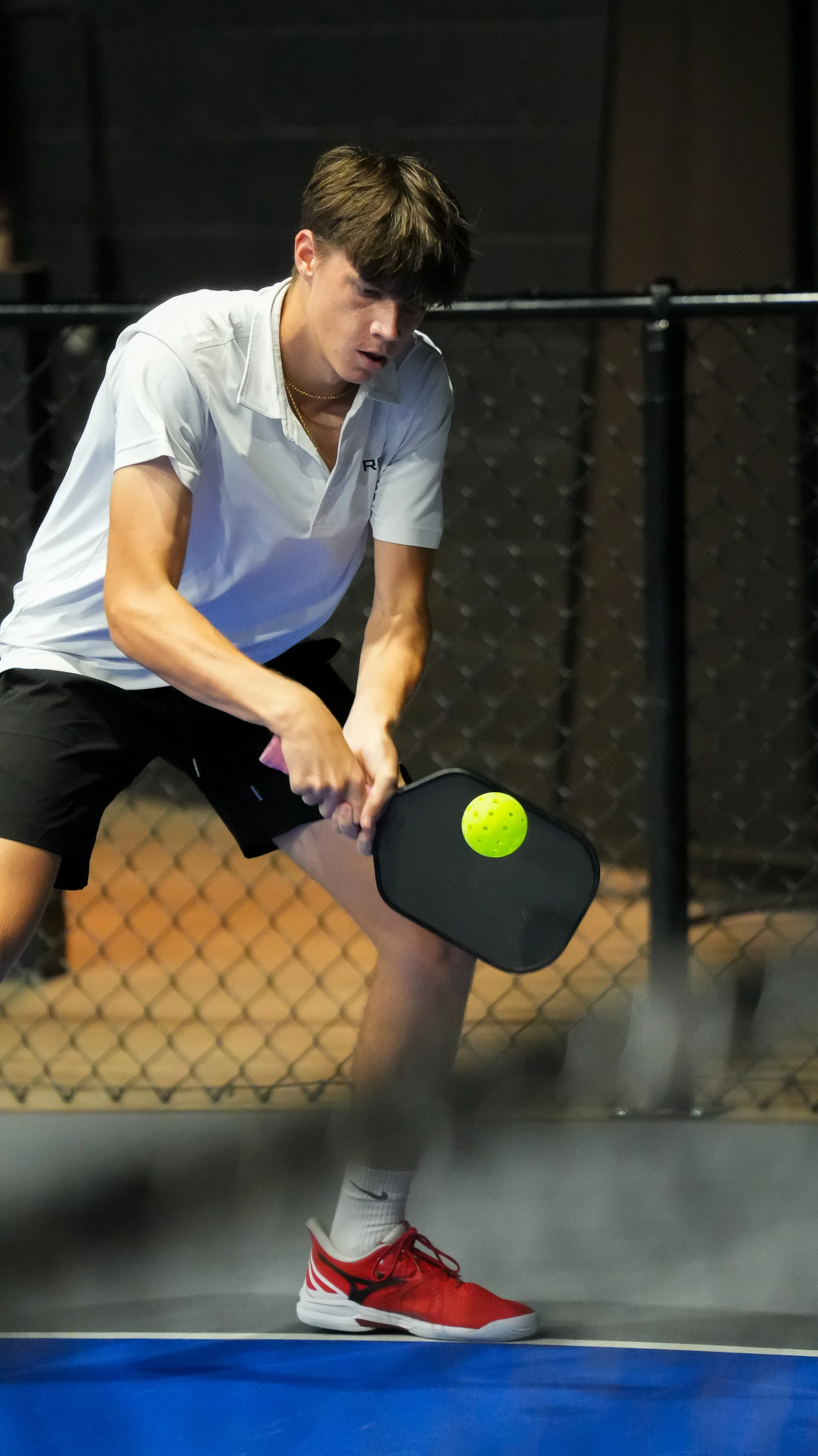 A young man playing pickleball, preparing to hit a yellow ball with a black paddle, wearing a white shirt, black shorts, white socks, and red sneakers on an indoor court with a chain-link fence in the background.