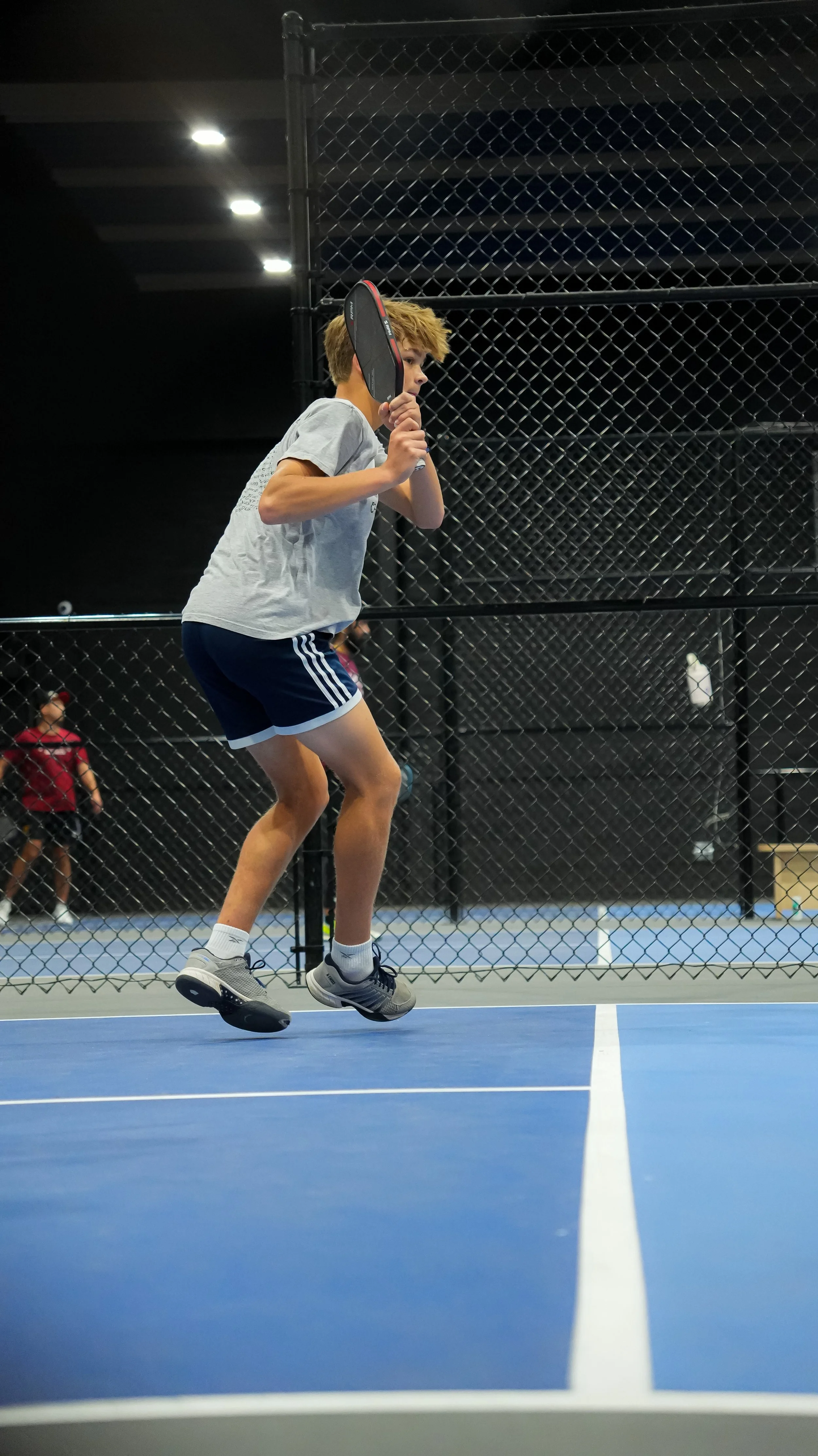 A young boy in sports attire playing tennis on an indoor court, jumping with a tennis racket near his face, with a black chain-link fence and other players in the background.