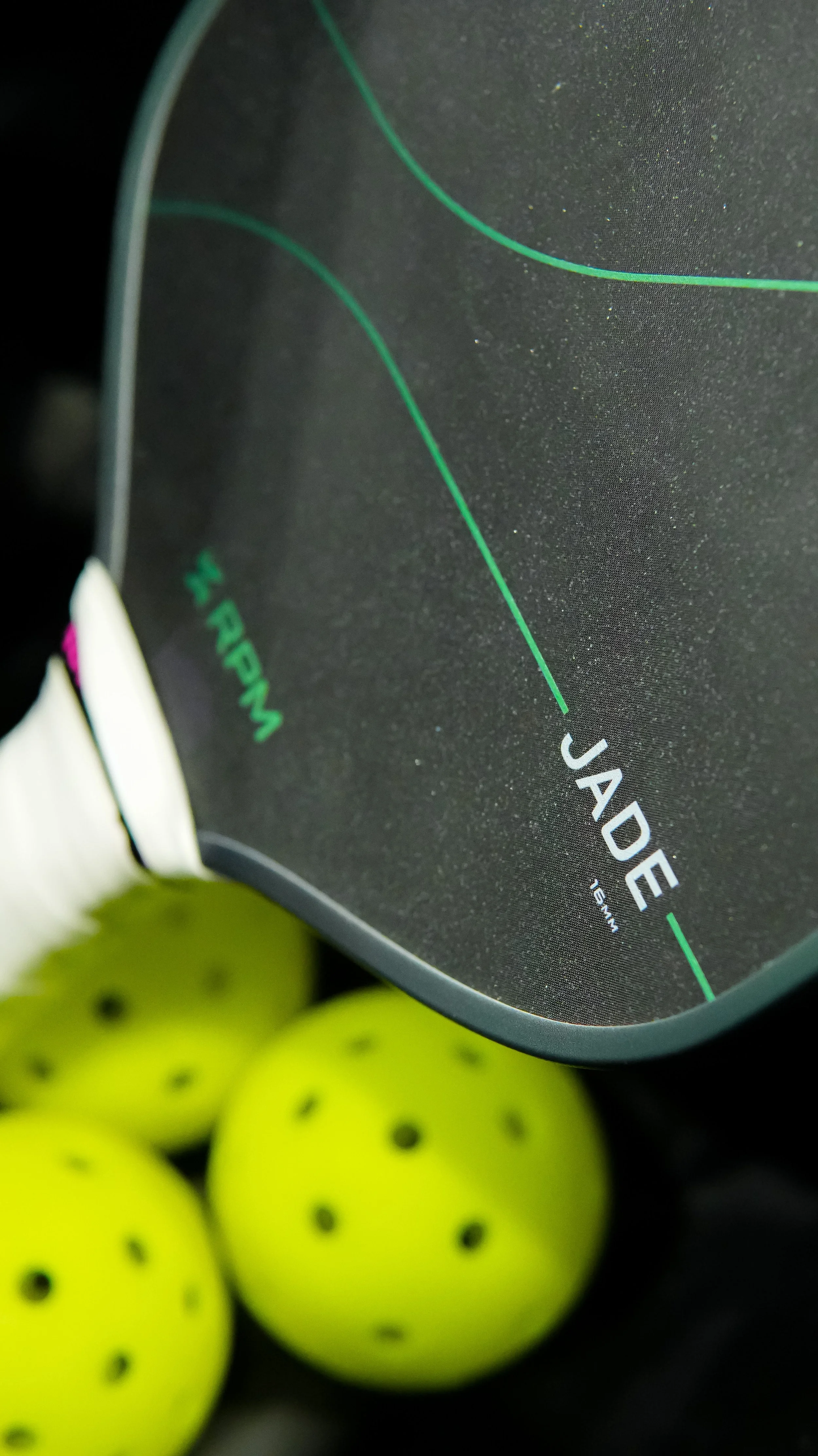 Close-up of a black tennis racket with green lines and the word 'Jade' printed on it, surrounded by yellow tennis balls with black dots.