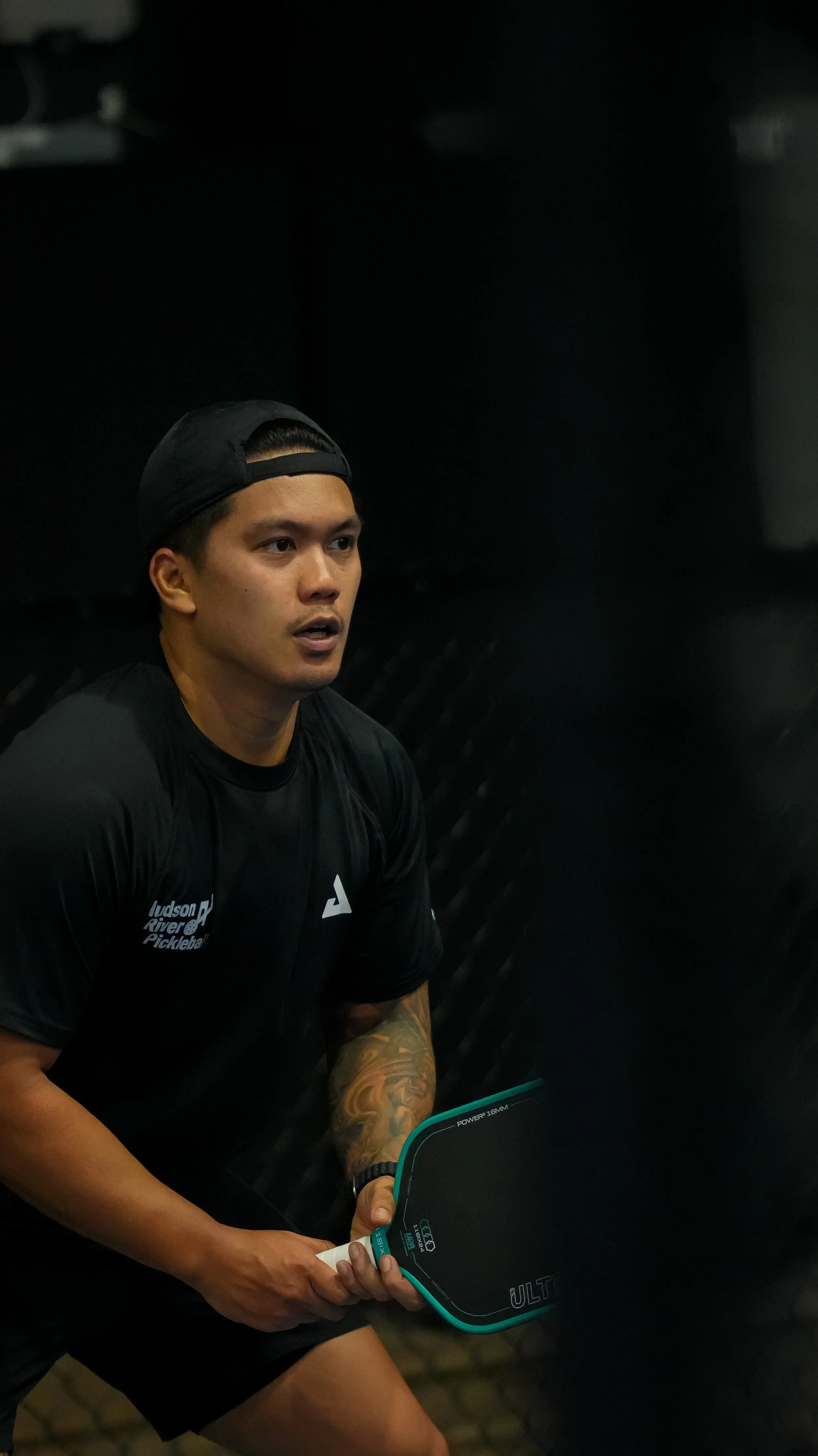 A young man wearing a black cap backwards, black athletic shirt with white logo, and black shorts sitting indoors, holding a tablet with various data displayed on the screen.