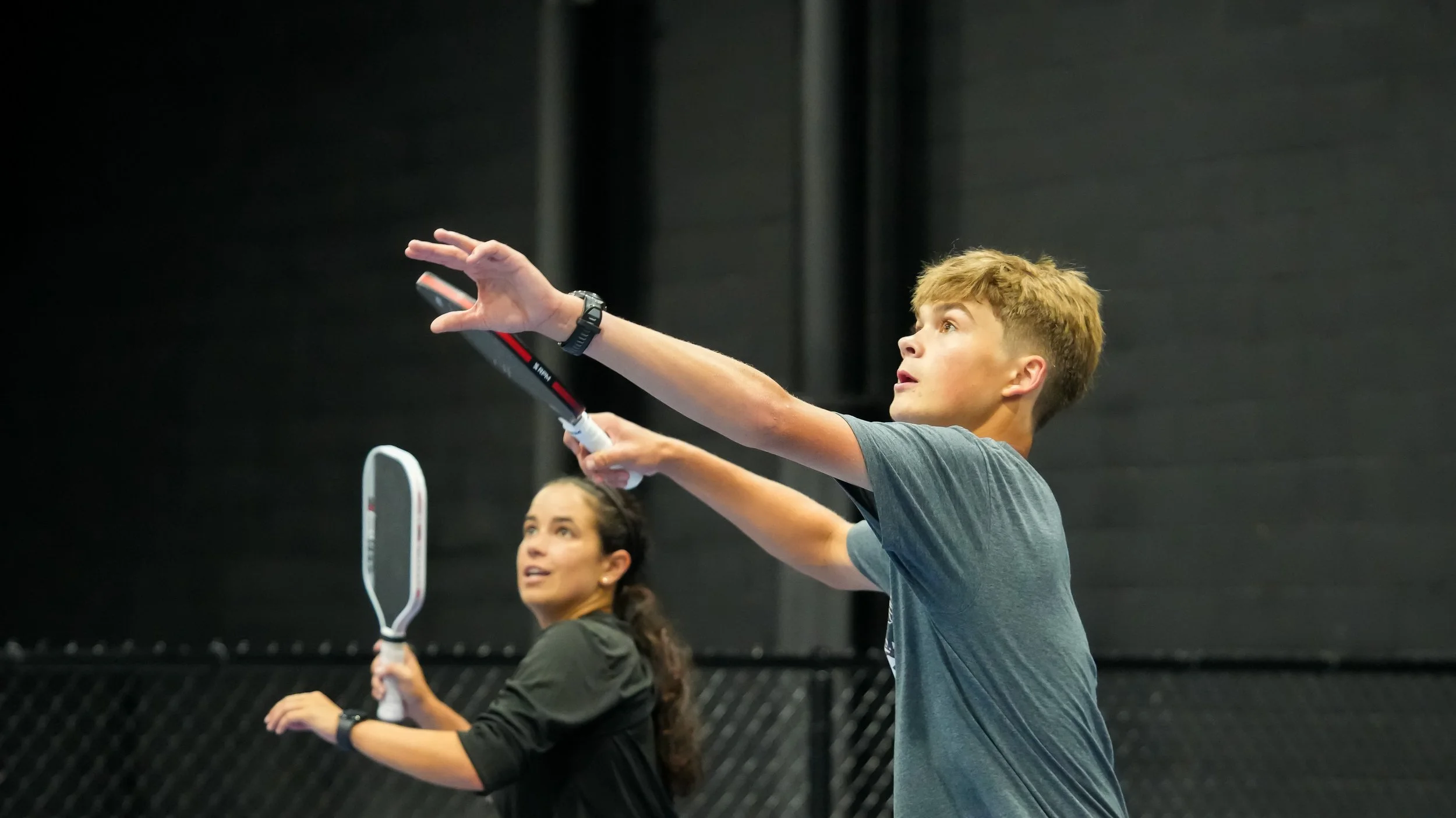 A young man and woman playing badminton indoors, both holding rackets and preparing to hit the shuttlecock.