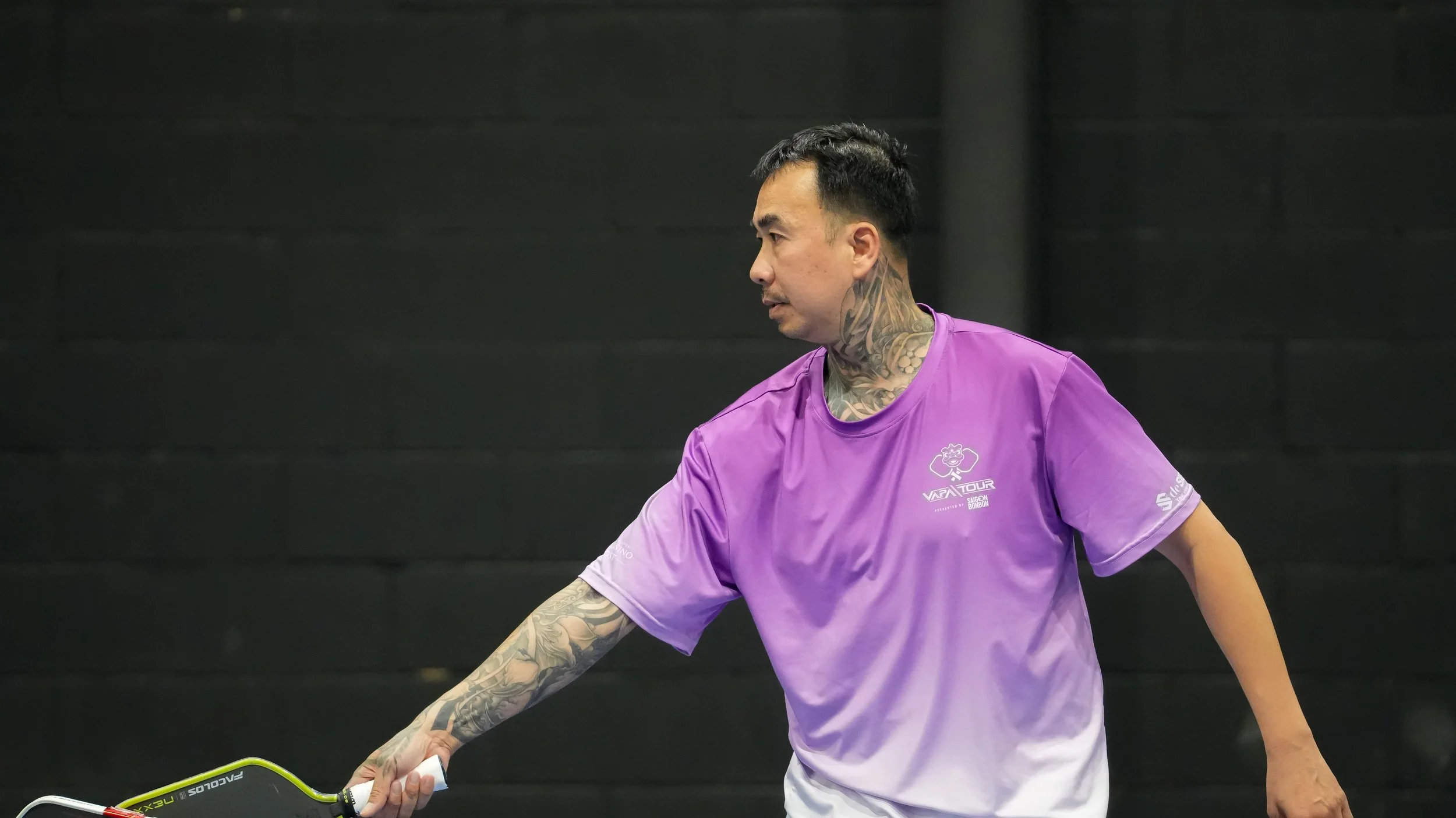 A man with tattoos on his neck and arms is playing tennis indoors, wearing a purple shirt with 'VAPATOUR' and other logos.