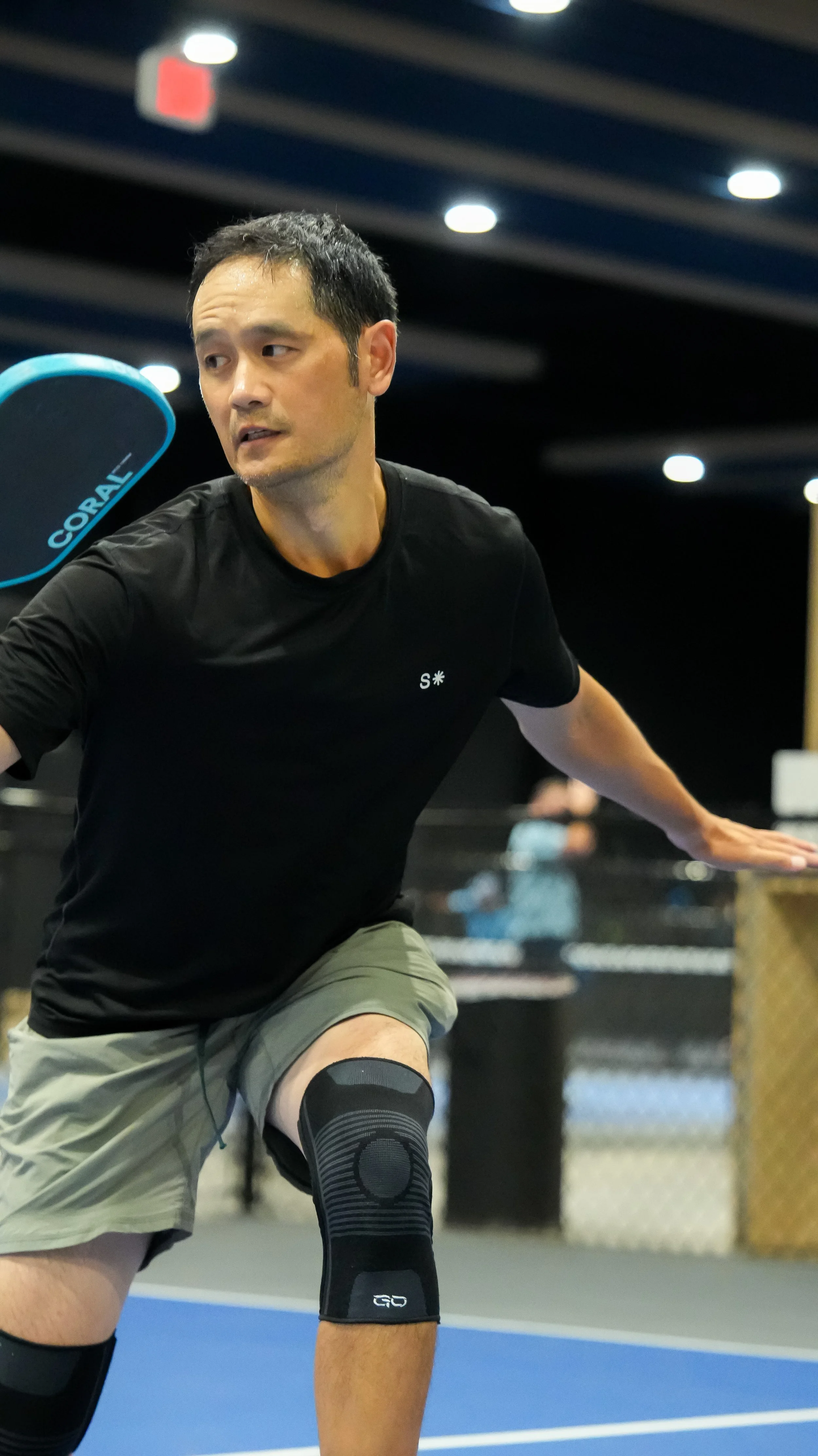 A man with short dark hair in a black shirt and gray shorts playing pickleball indoors. He's wearing a black knee brace and black compression socks, with a blue pickleball paddle in the background.
