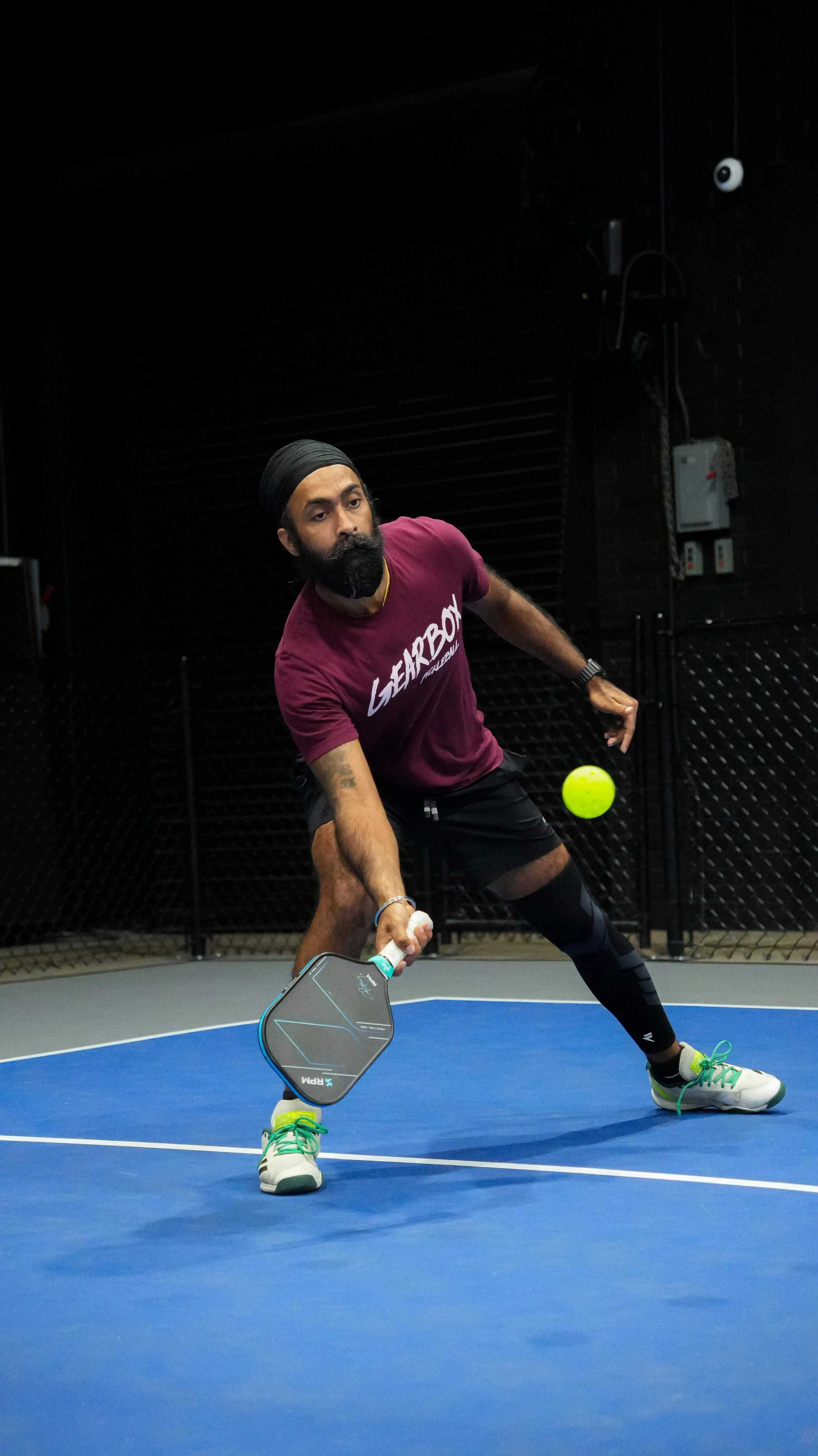 A man playing tennis indoors on a blue court, dressed in a maroon t-shirt, black shorts, and white tennis shoes with green laces, preparing to hit a tennis ball with a paddle.