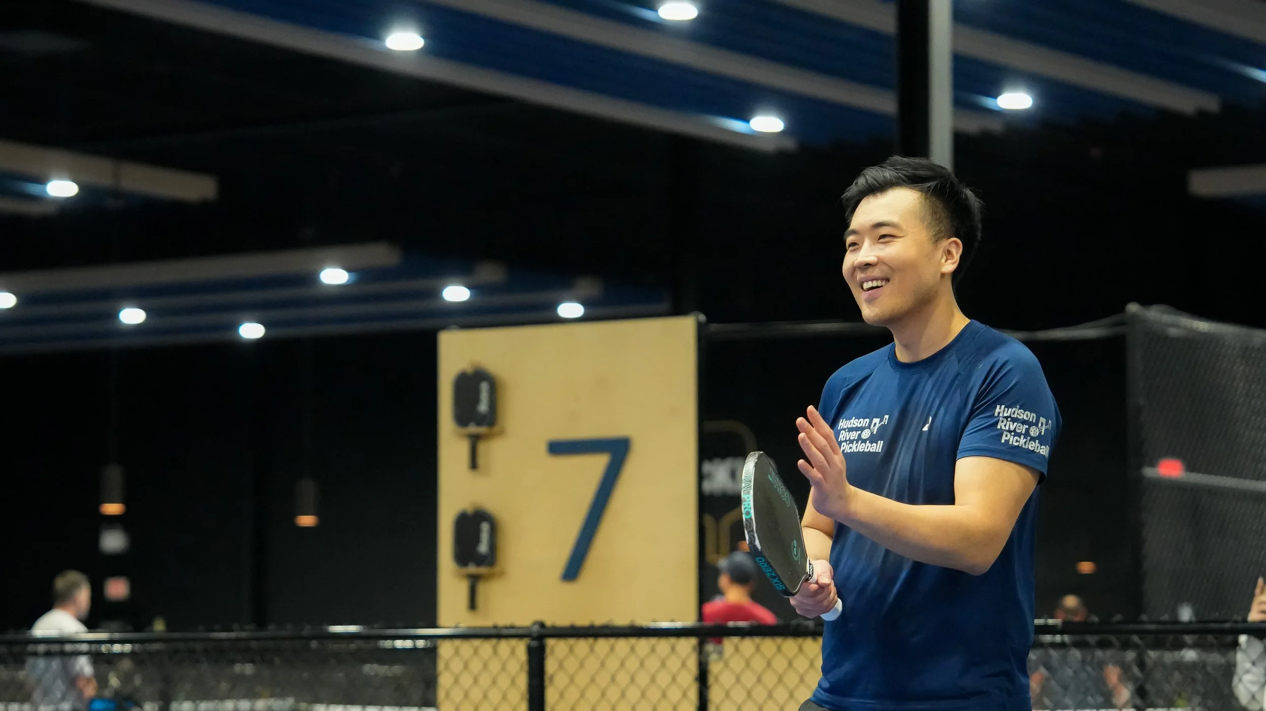 A man smiling while holding a pickleball paddle in a gym with blue ceiling and yellow partition. He is wearing a blue T-shirt with white text that reads 'Hudson River Pickleball'.