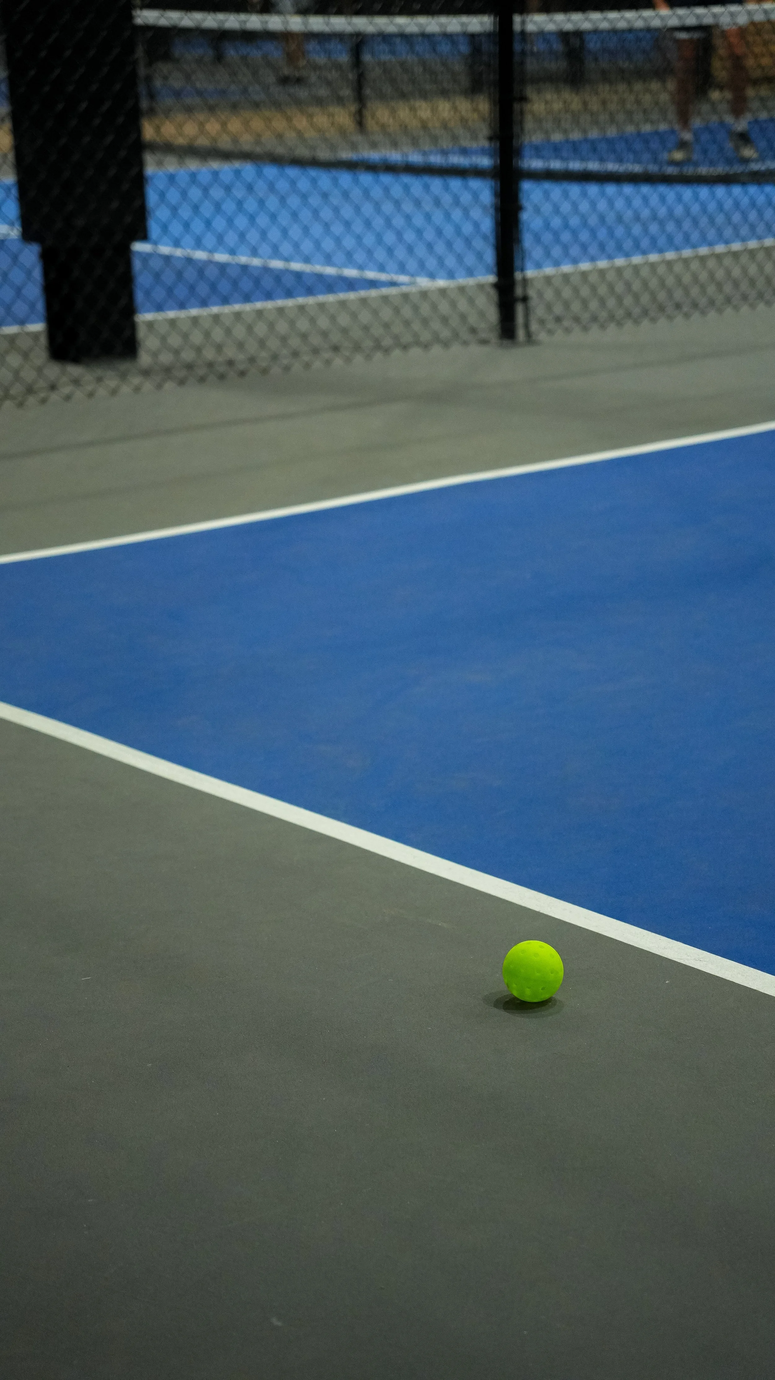An outdoor tennis court with a bright green tennis ball on the dark surface, and a blue tennis court visible in the background behind a chain-link fence.