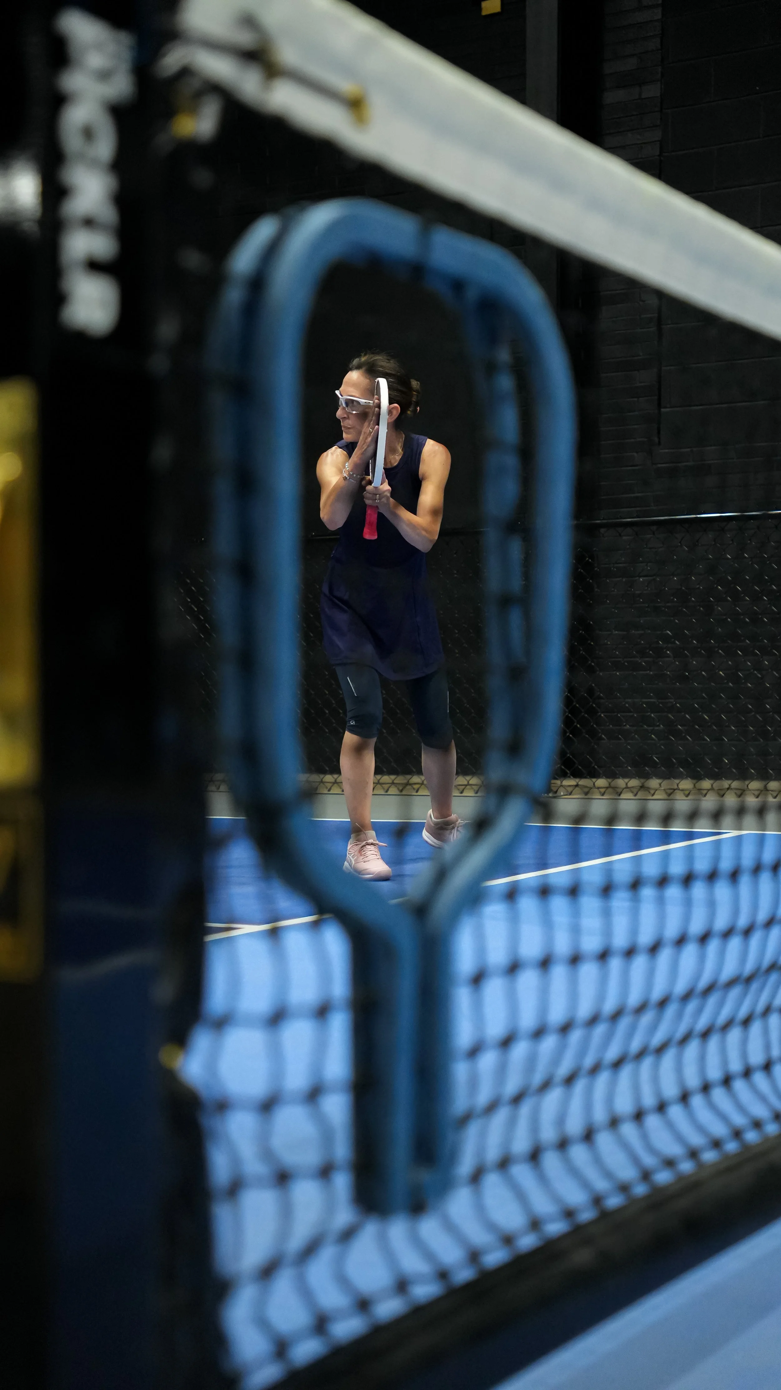 A woman wearing sportswear, goggles, and gloves standing on a tennis court, holding a tennis racket, viewed through a chain-link fence with a blue batting cage in the foreground.