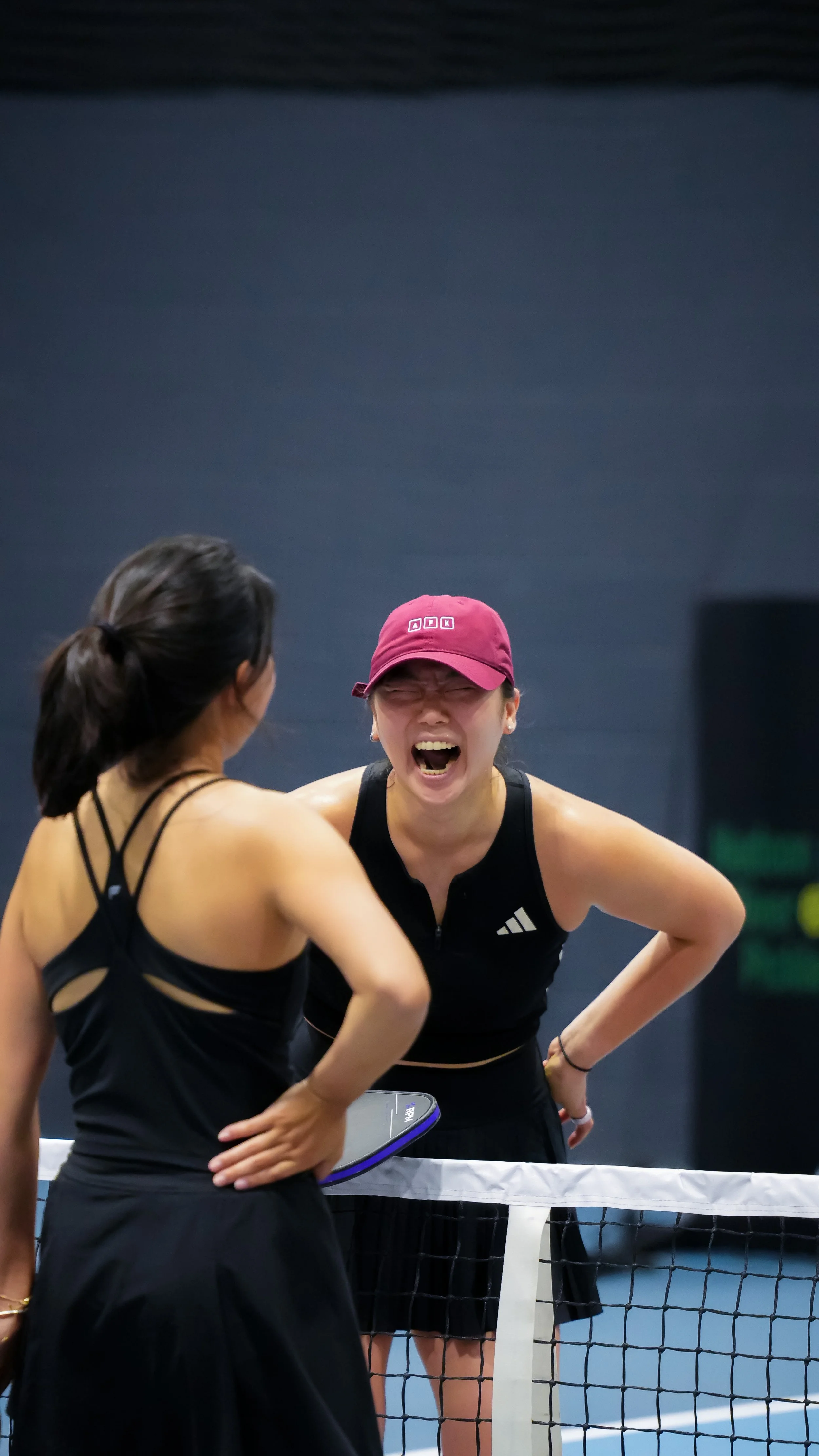 Two women with athletic clothing and a ping pong paddle are having a serious conversation at a ping pong table, with one woman appearing emotional.
