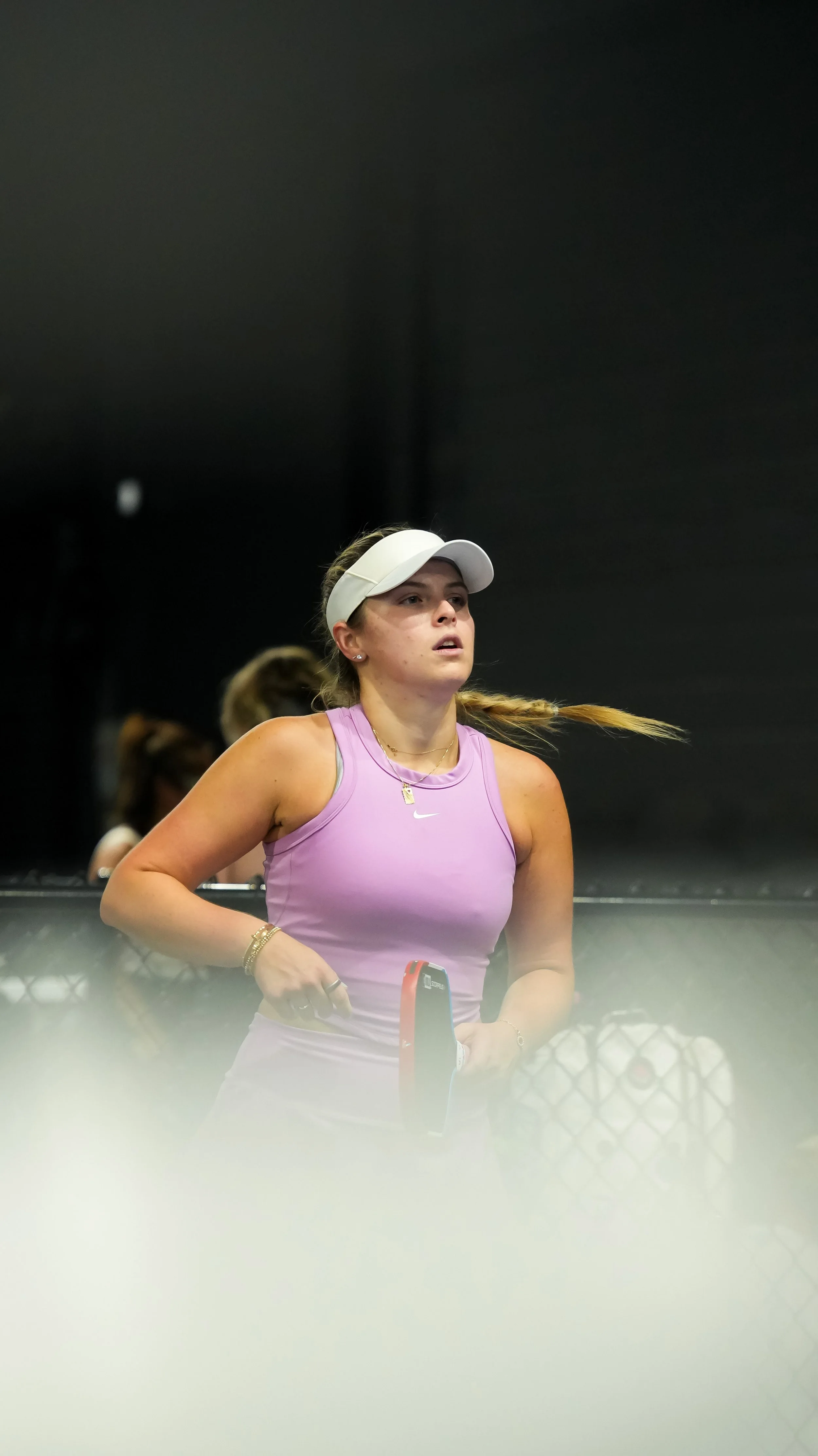 A woman wearing a white visor and pink sports top standing behind a tennis net with a tennis racket in hand, on a tennis court.