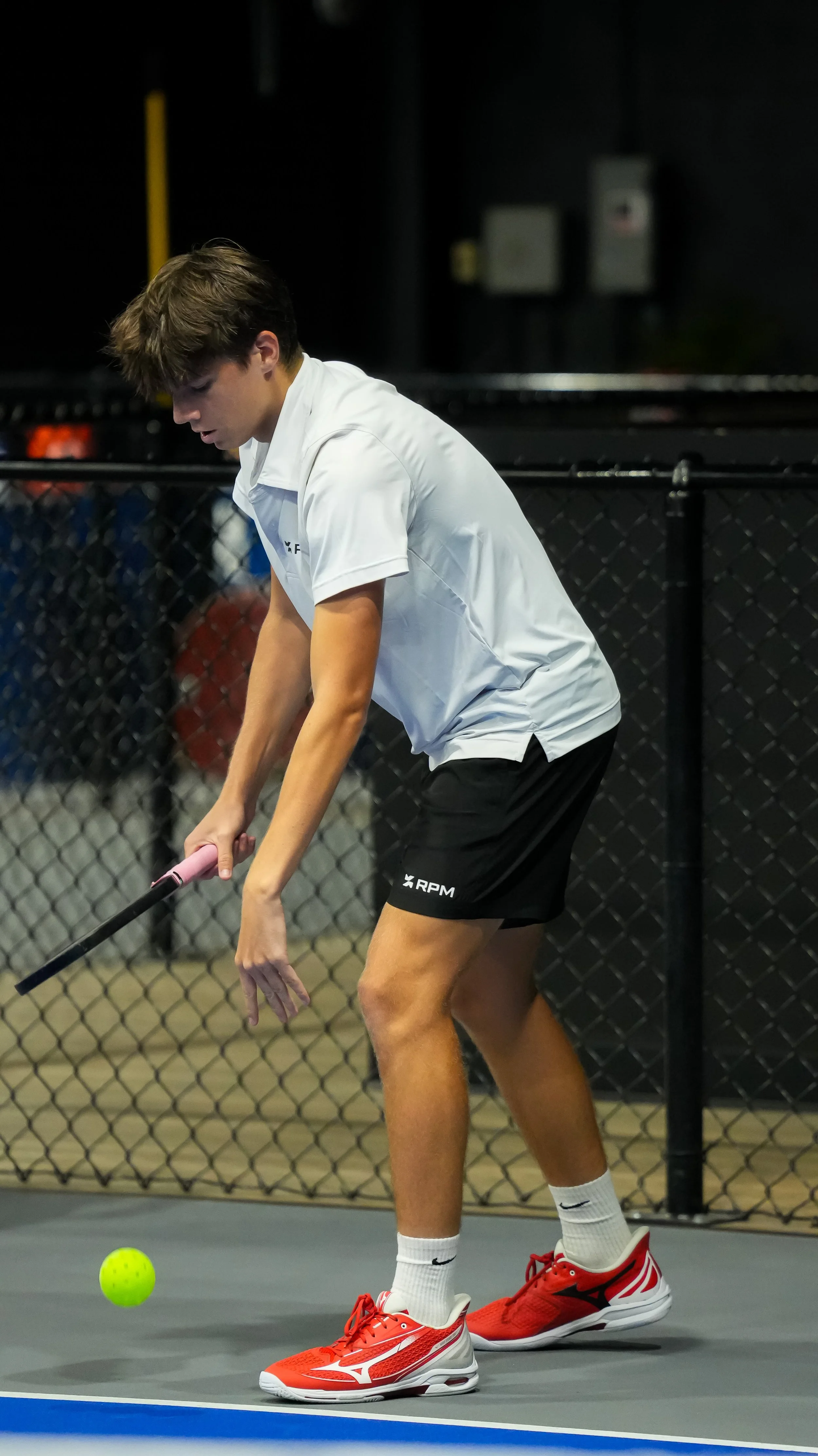 A young man in a white polo shirt, black shorts, and red tennis shoes practicing tennis indoors. He is holding a tennis racket and concentrating on a bright yellow tennis ball near his feet, with a black chain-link fence in the background.