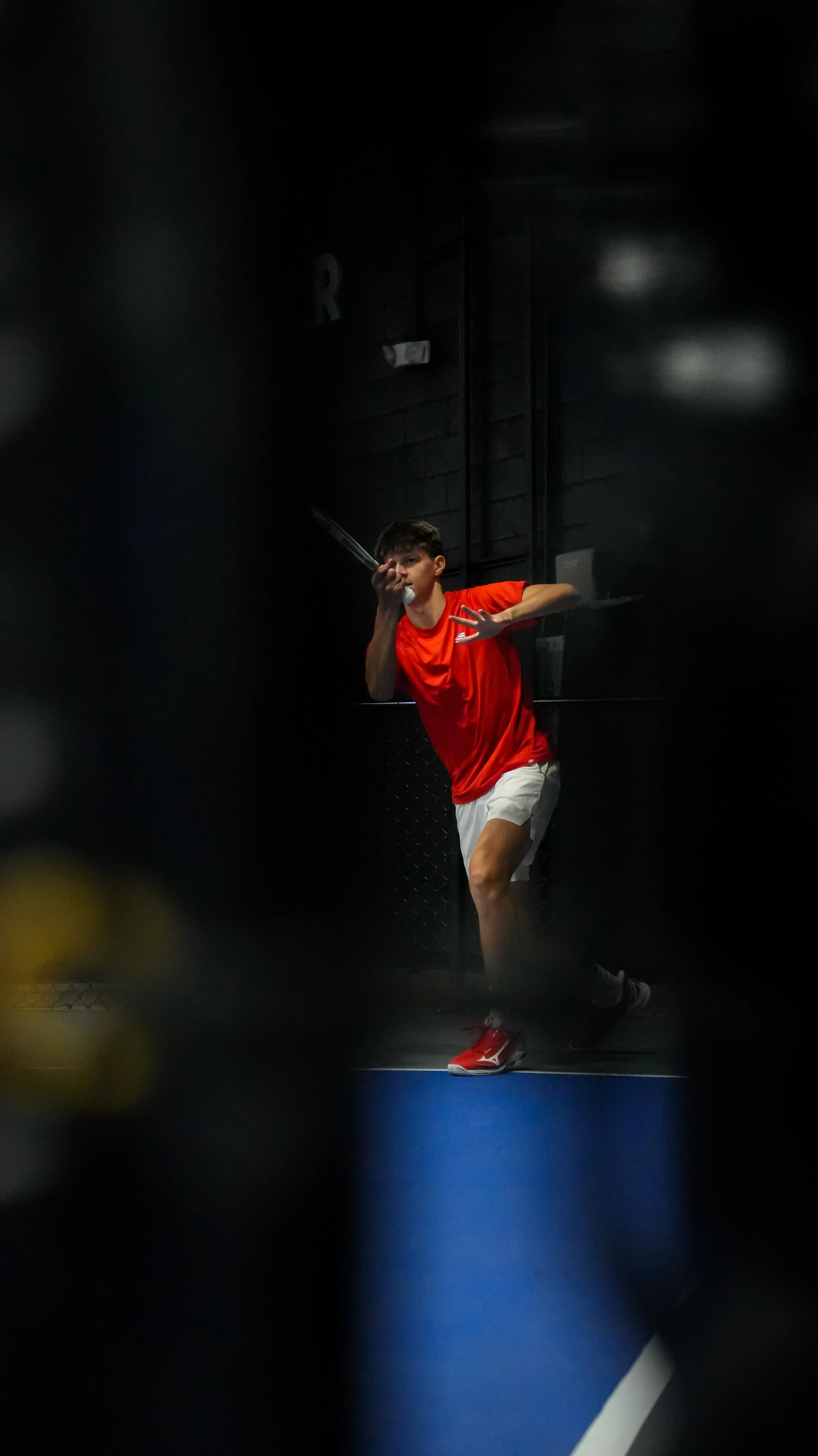 A young man in a red athletic shirt and white shorts playing ping pong in a dimly lit indoor gym or sports facility.