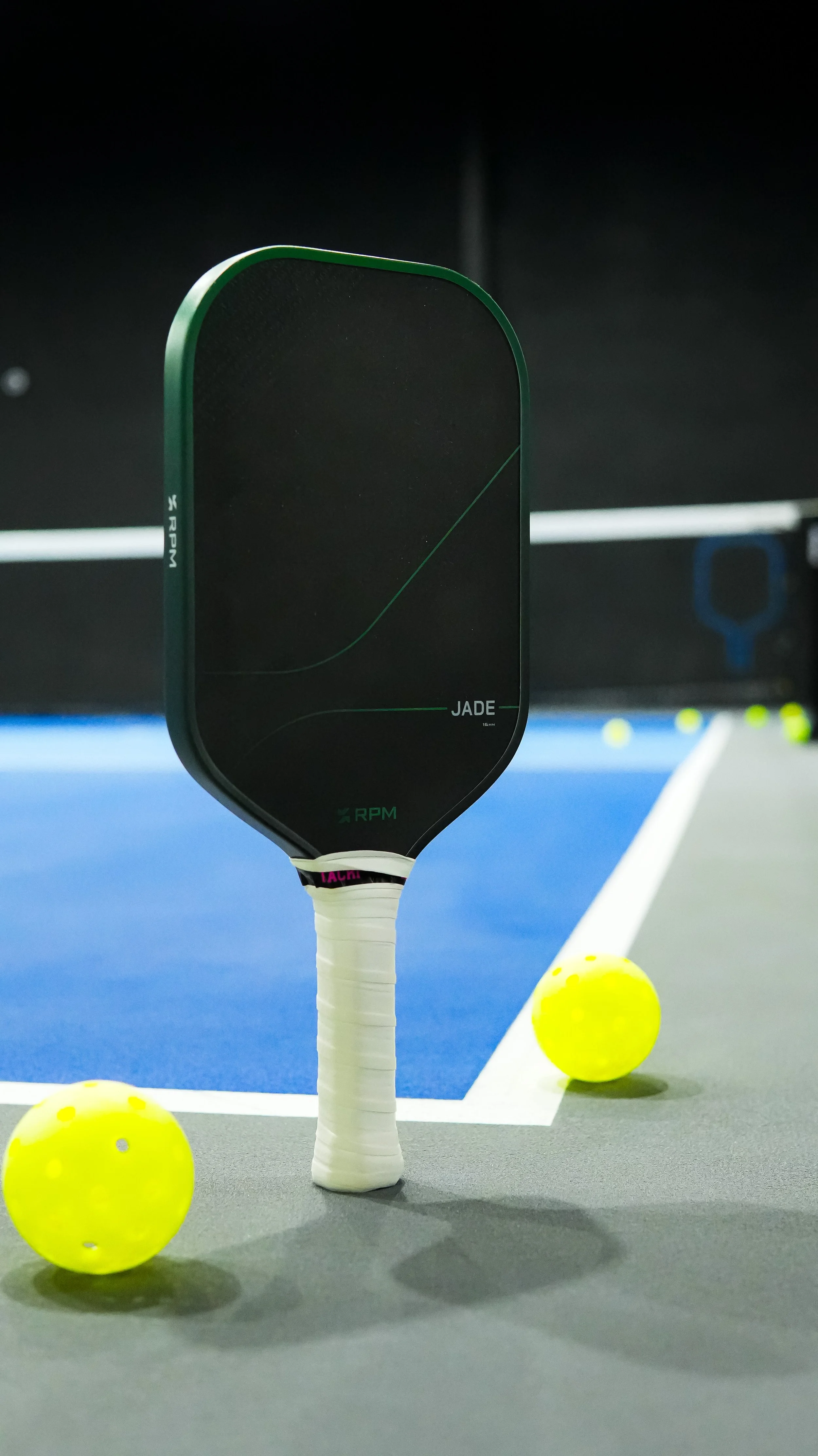 A pickleball paddle with a black face and green edges is standing upright on a gray court, with a yellow pickleball in front and a bright yellow ball and blue lines in the background.