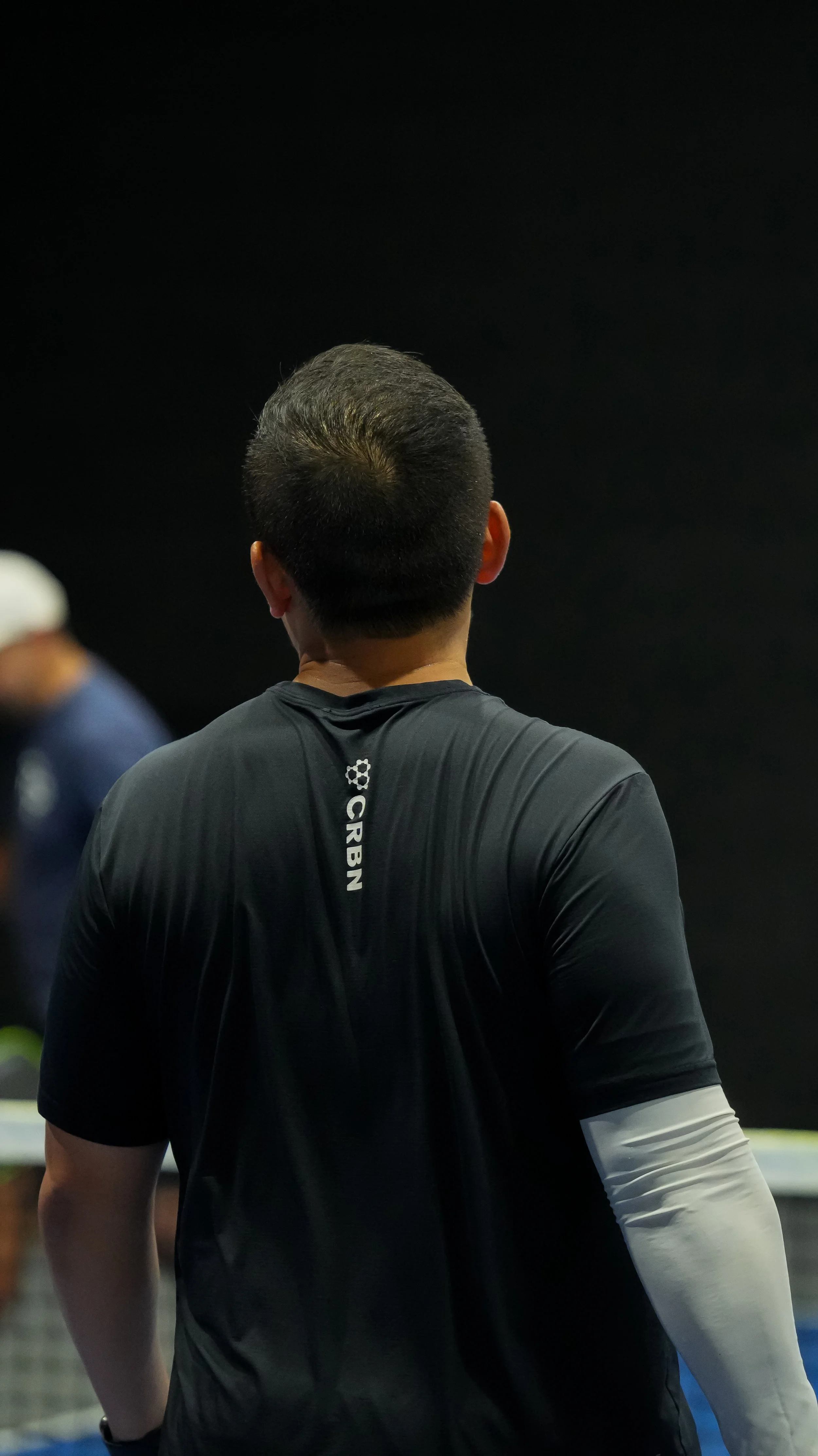 Back view of a man wearing a black shirt with white and blue details, standing in an indoor sports facility.