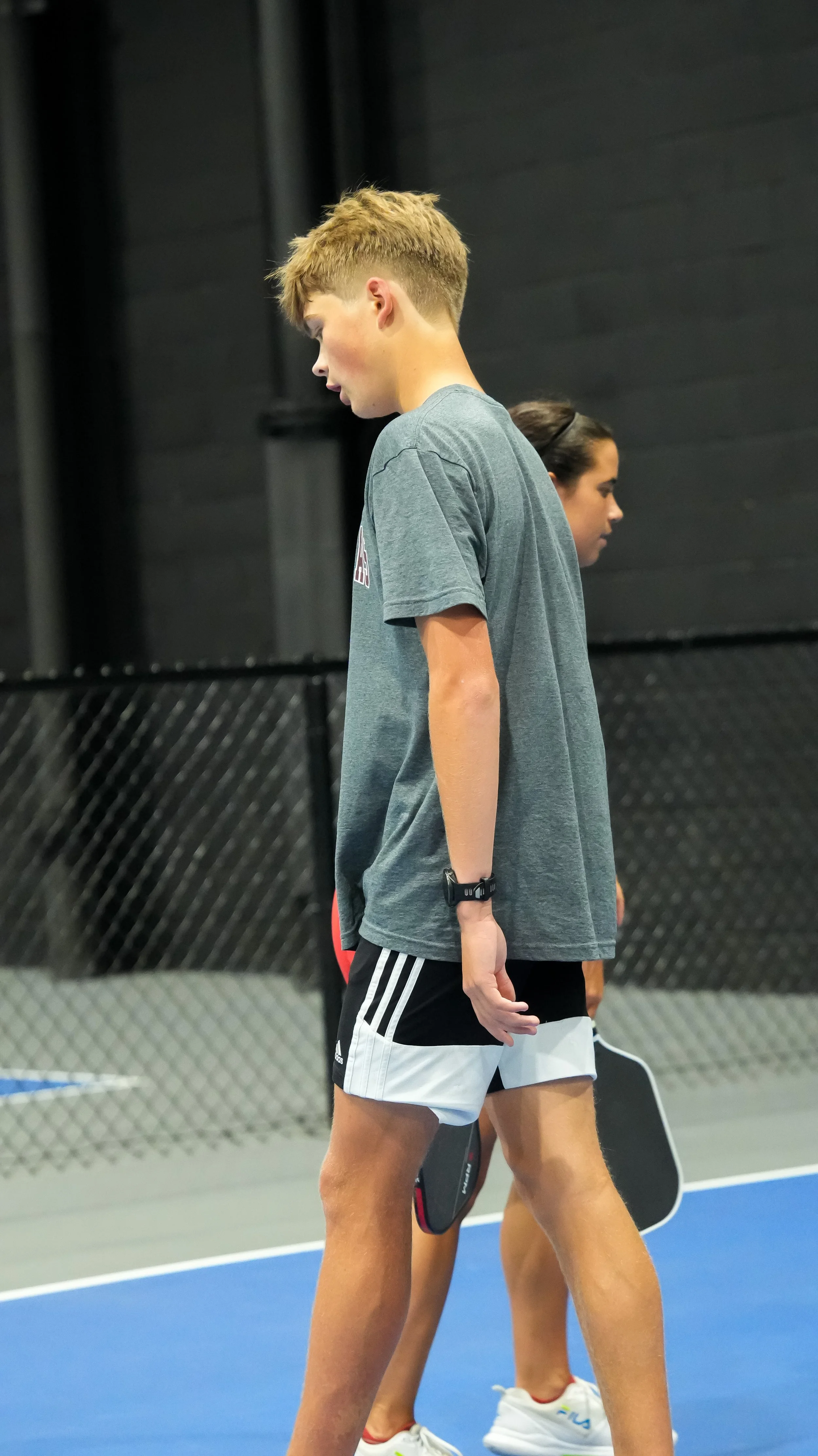 Two young athletes standing on a blue indoor sports court, holding paddle tennis paddles, preparing for a game.