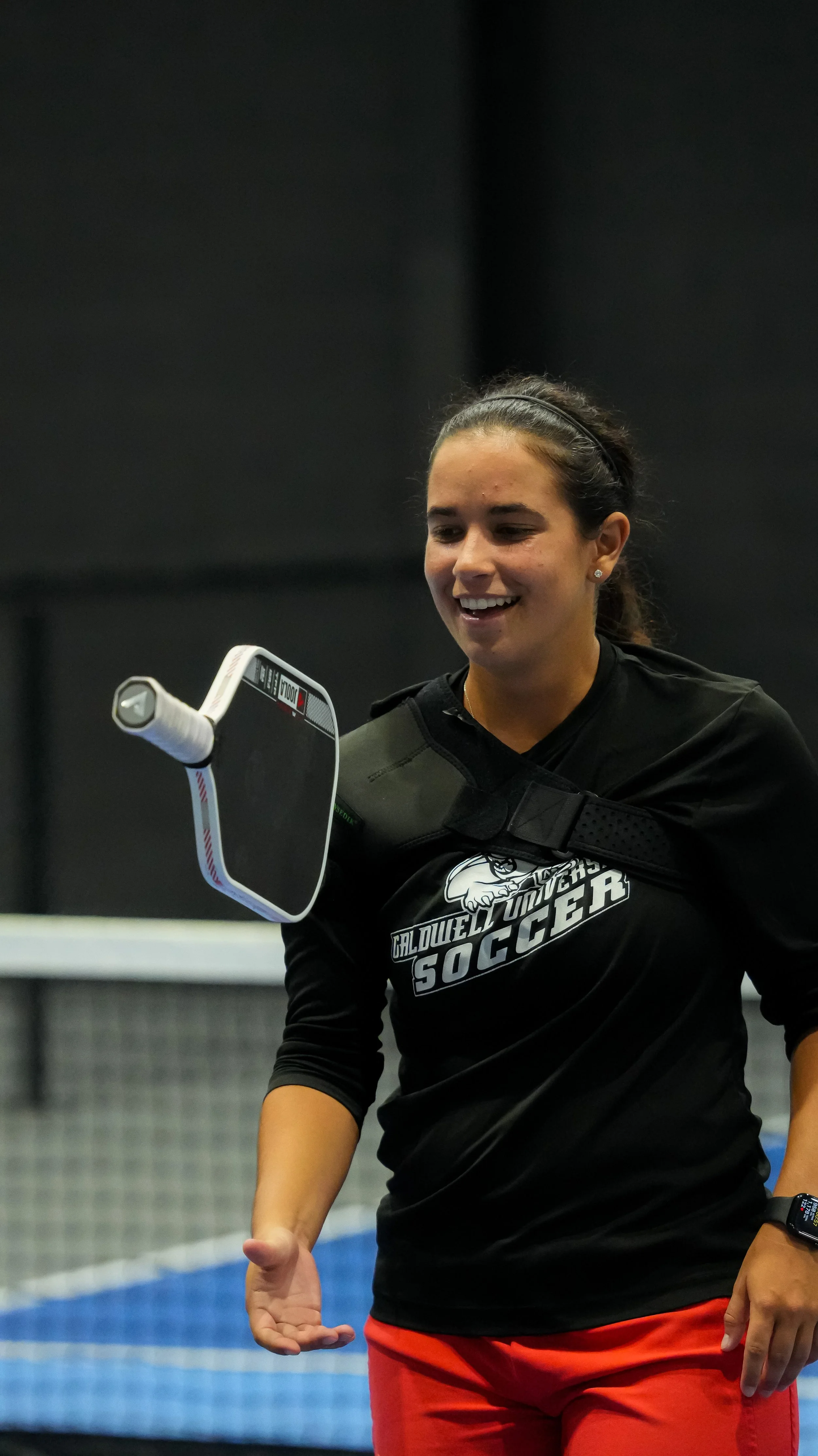 A young woman with dark hair tied back, wearing a black Caldwell University soccer shirt, is smiling and looking at a cushioned training device hanging in front of her. She is in a sports facility with blue and gray walls.