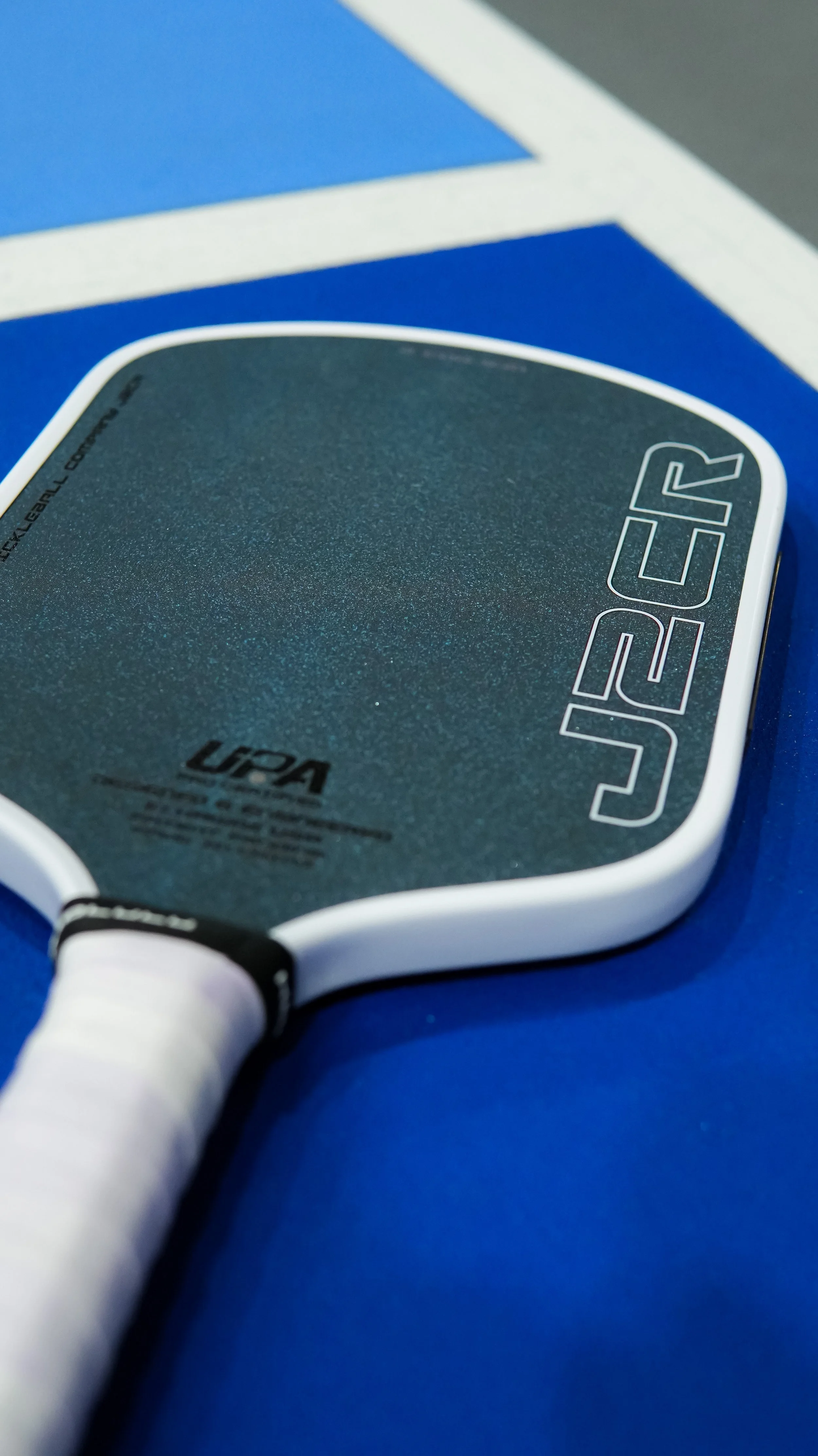 Close-up of a table tennis paddle with a black rubber surface and white handle, resting on a coarse blue surface.