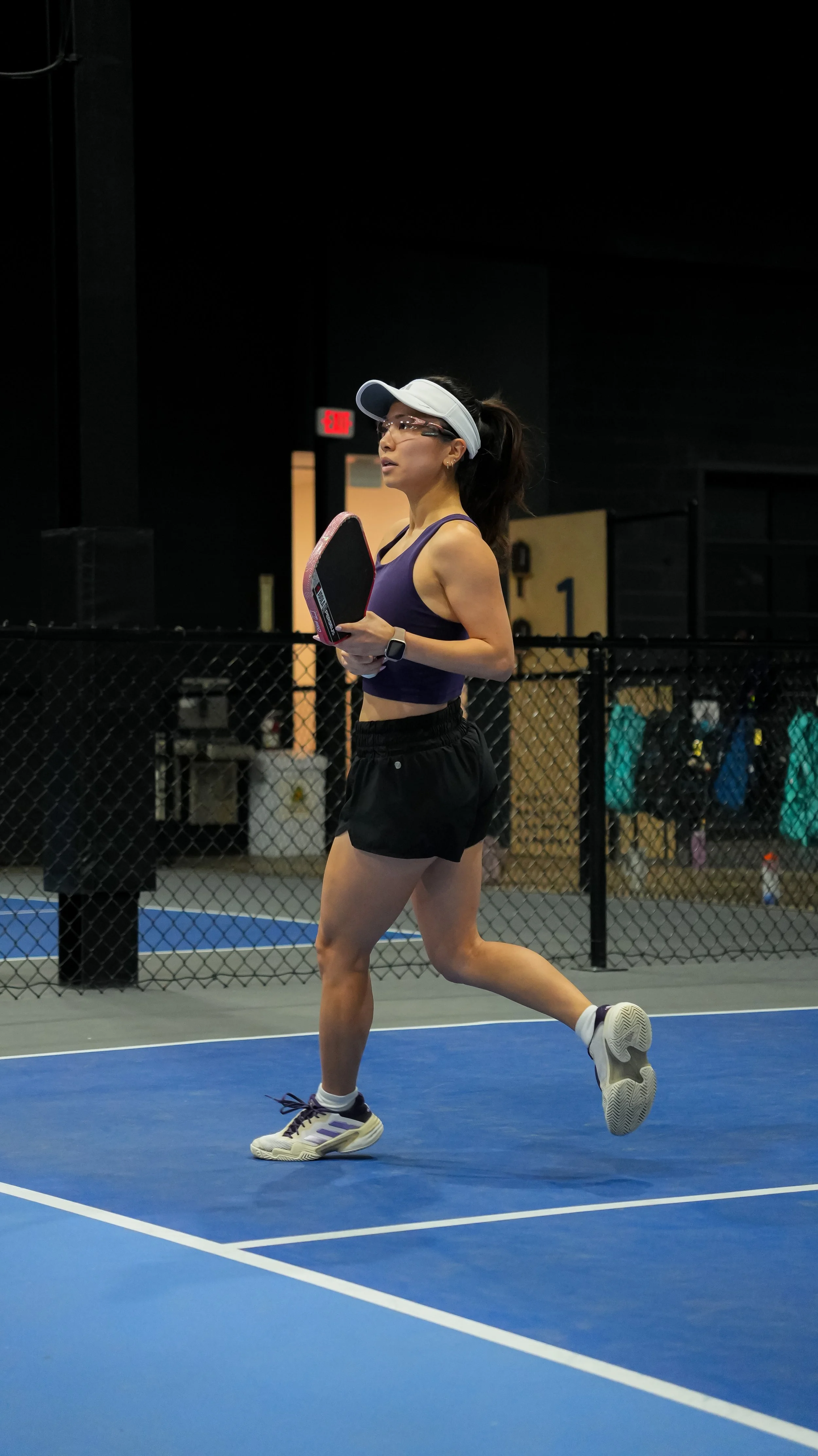 A woman in athletic clothes running on an indoor tennis court, holding a pickleball paddle, with a black fence and gym bags in the background.