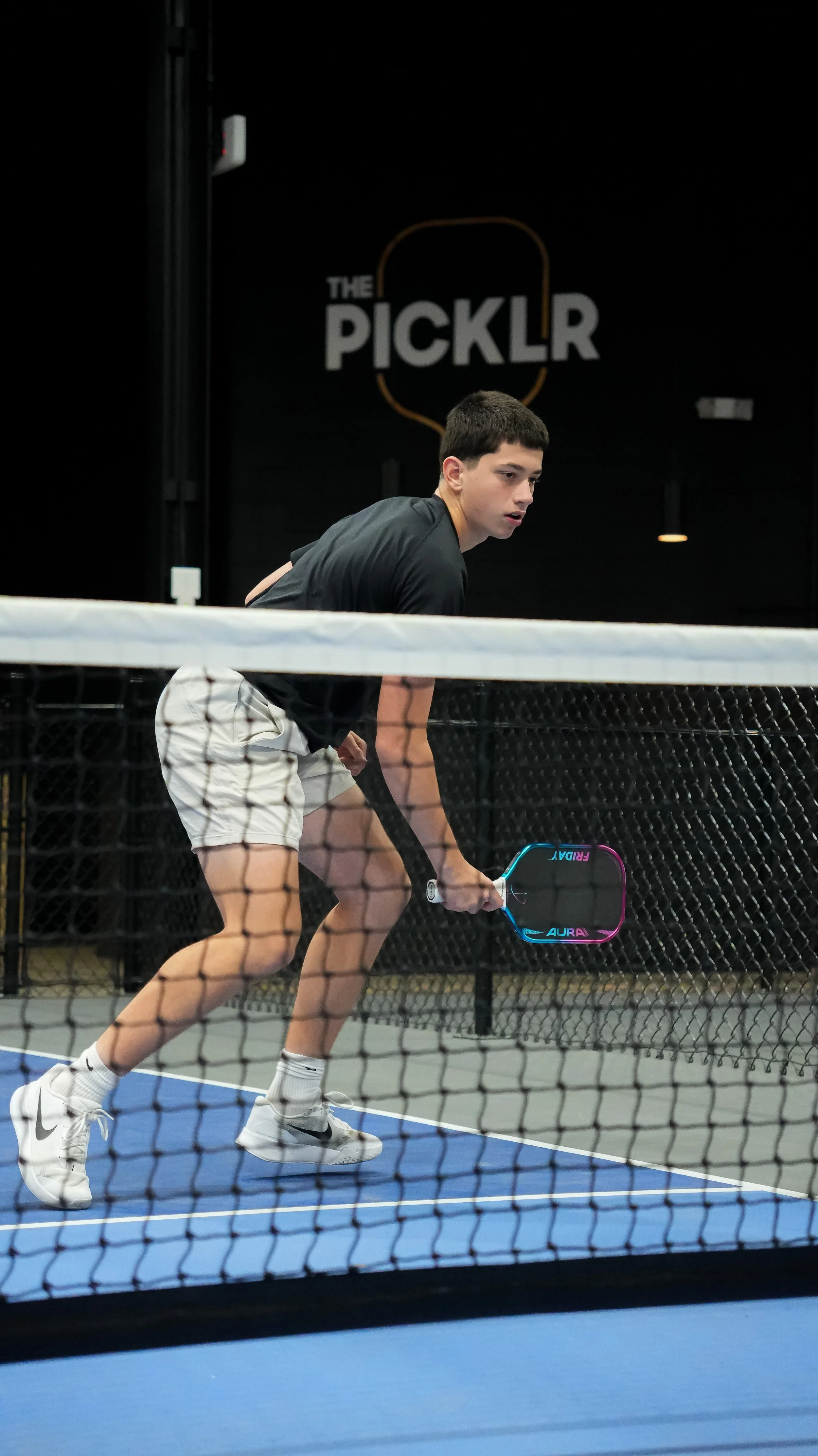 A young man playing pickleball on an indoor court, holding a paddle and preparing to hit the ball. In the background, there is a sign that reads 'The Picklr'.