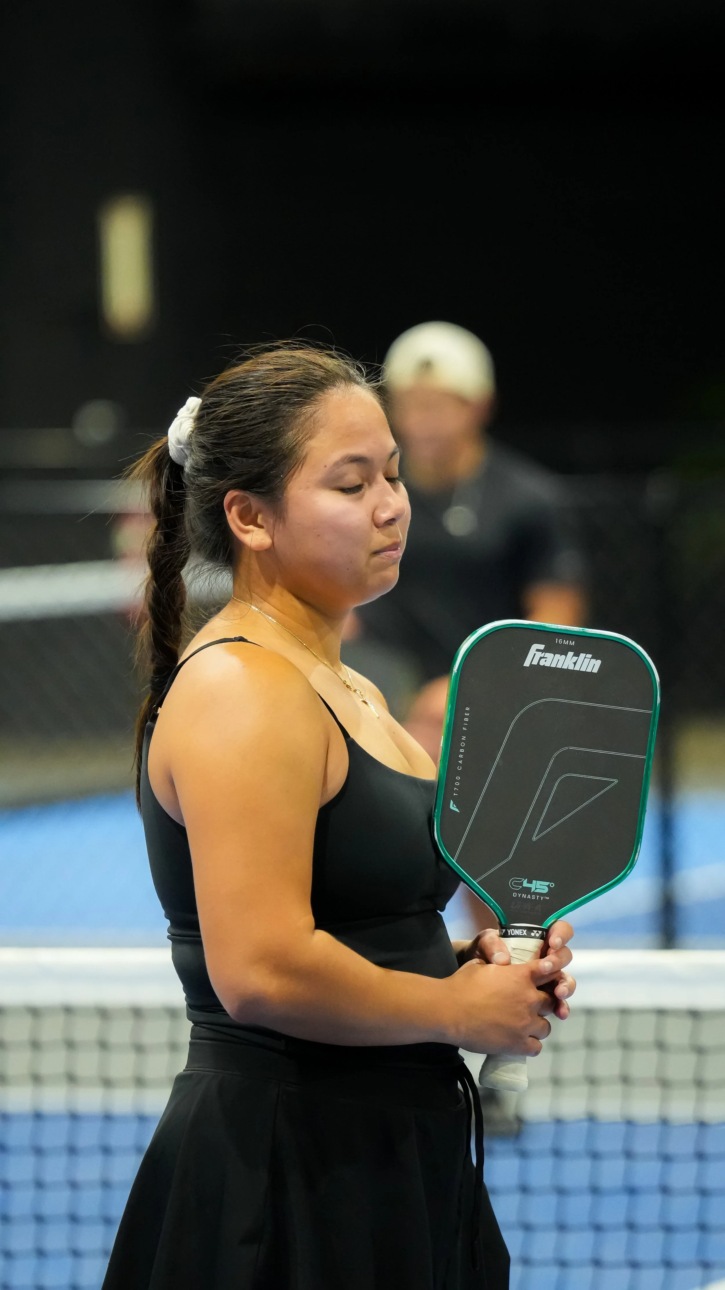A woman in a black sports top holding a tennis racket on a tennis court, with a man wearing a cap blurred in the background.