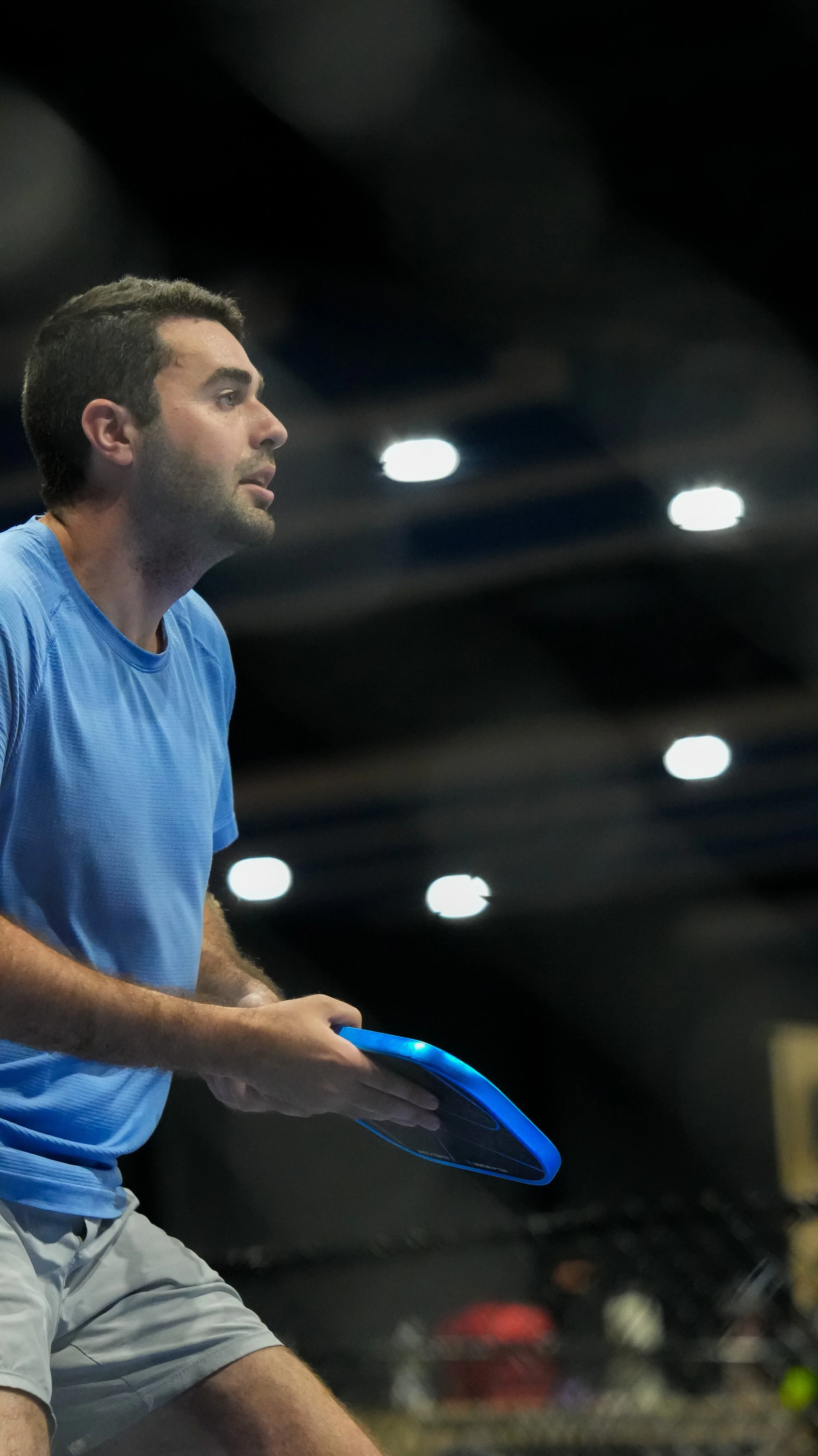 A man in a blue shirt and light-colored shorts holding a blue electronic tennis racket, likely during a match or practice, in an indoor setting with lights on the ceiling.
