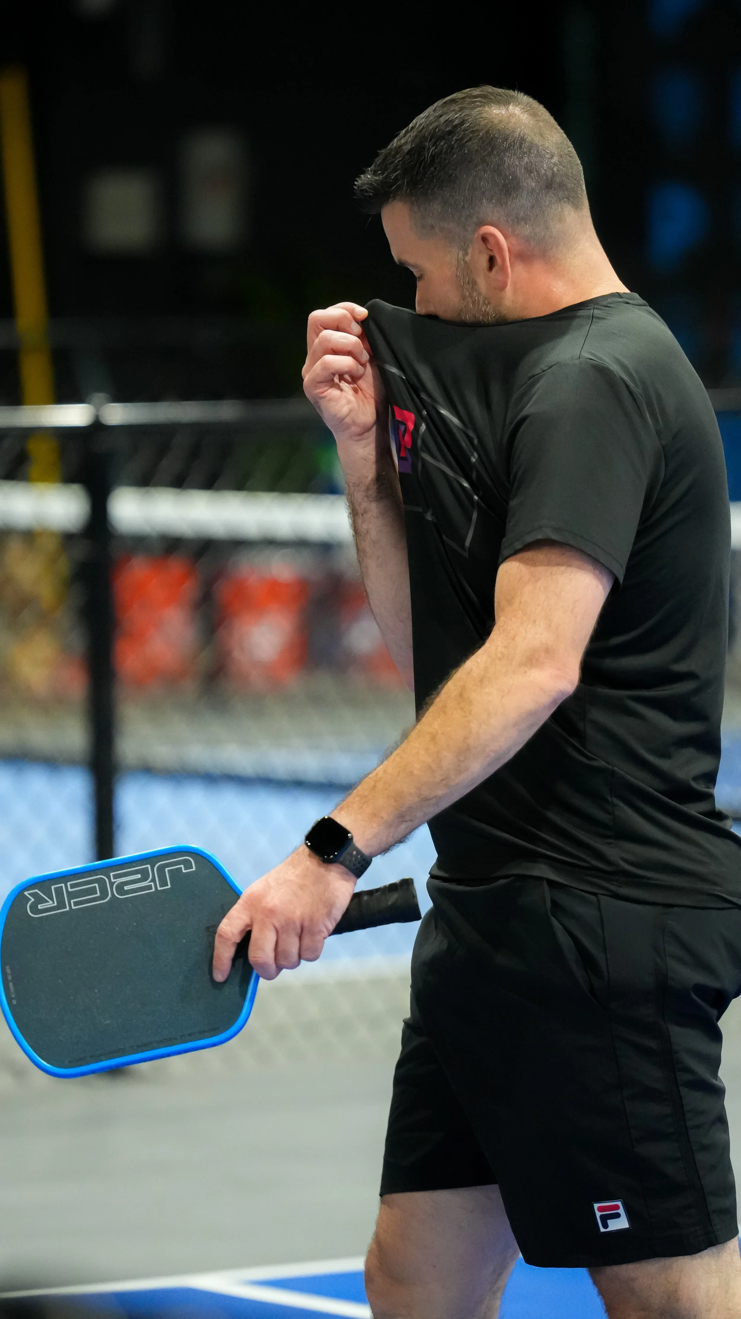 A man in black sportswear is holding a paddle and sporting a watch, likely playing pickleball or tennis, on an outdoor court at night.