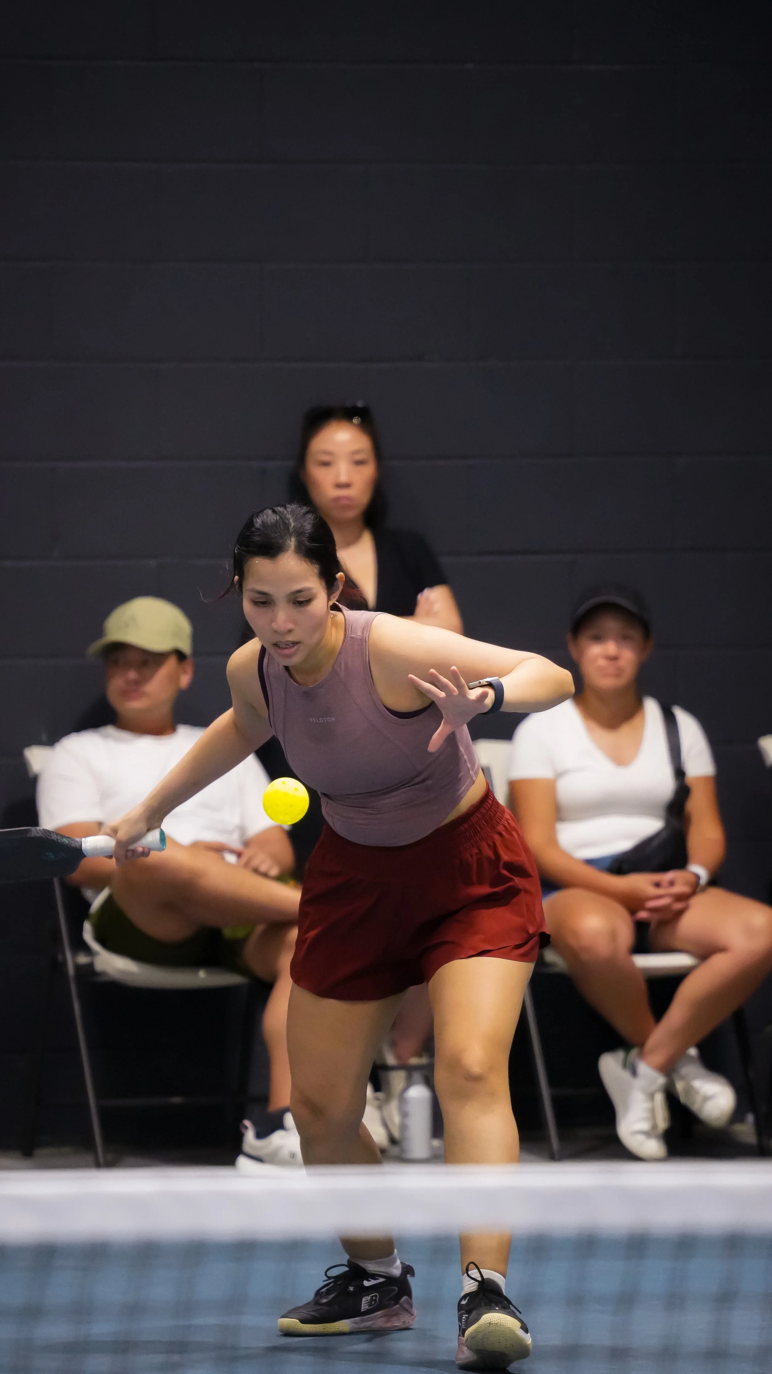 A woman in sportswear playing tennis or pickleball indoors, with three seated spectators watching behind her.