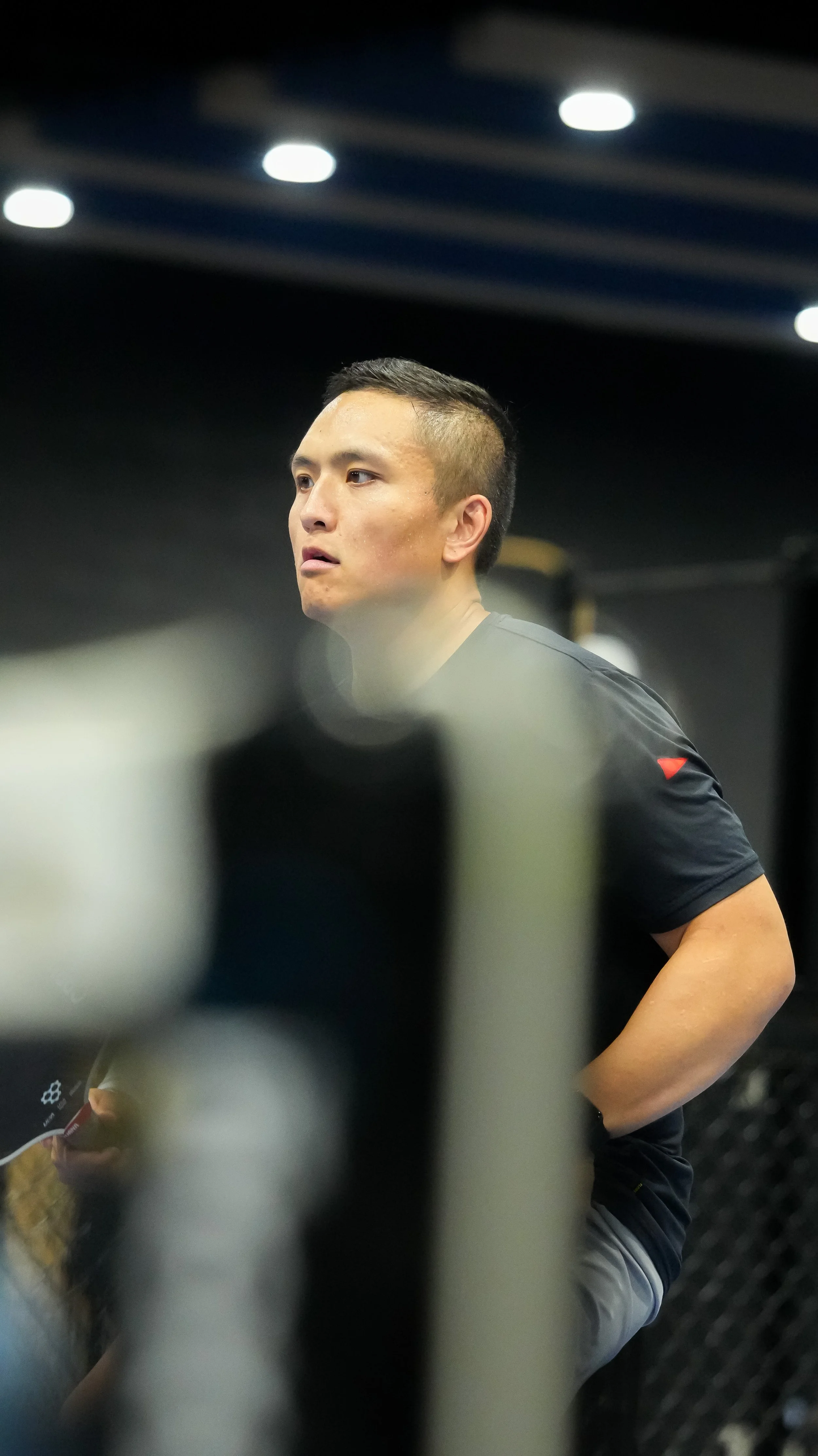 A man in a black athletic shirt with a red logo on the sleeve, looking to the left, in an indoor setting with ceiling lights.