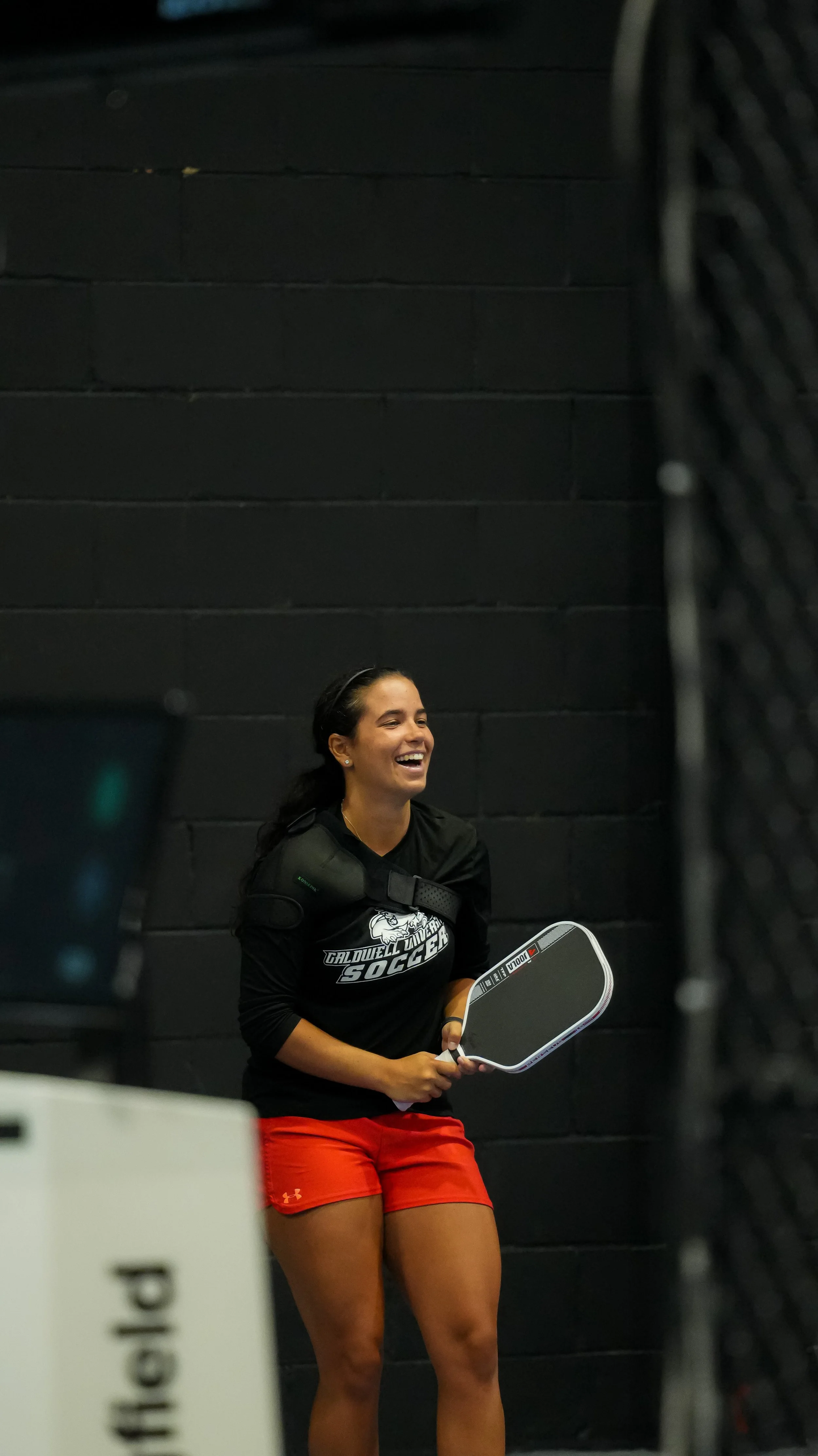 A woman in a black sweatshirt with 'Caldwell University Soccer' written on it and red shorts, smiling and holding a hockey stick, sitting against a black wall.