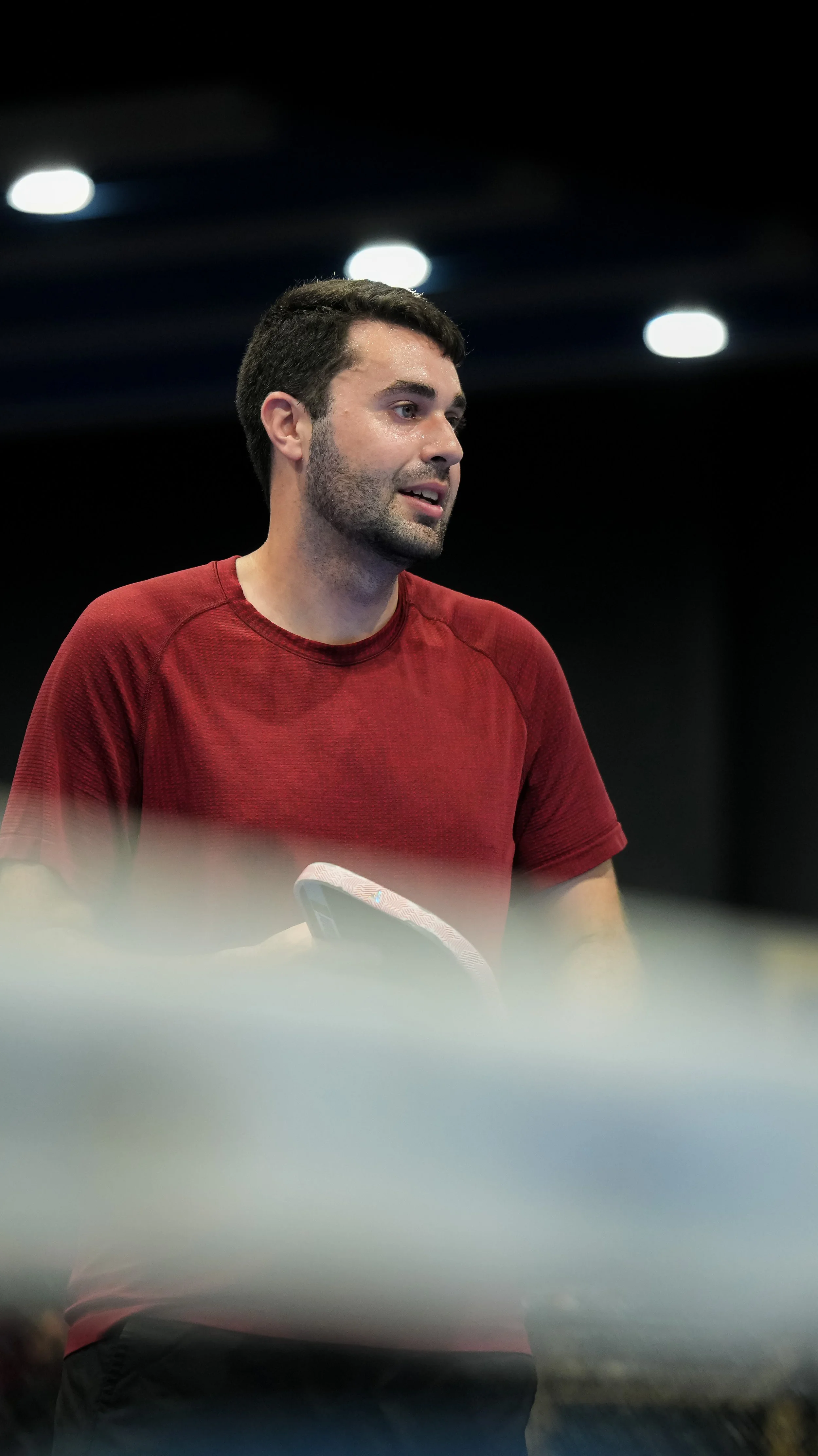 A man with dark hair and a beard wearing a red T-shirt and black pants, standing indoors with black ceiling and bright ceiling lights.
