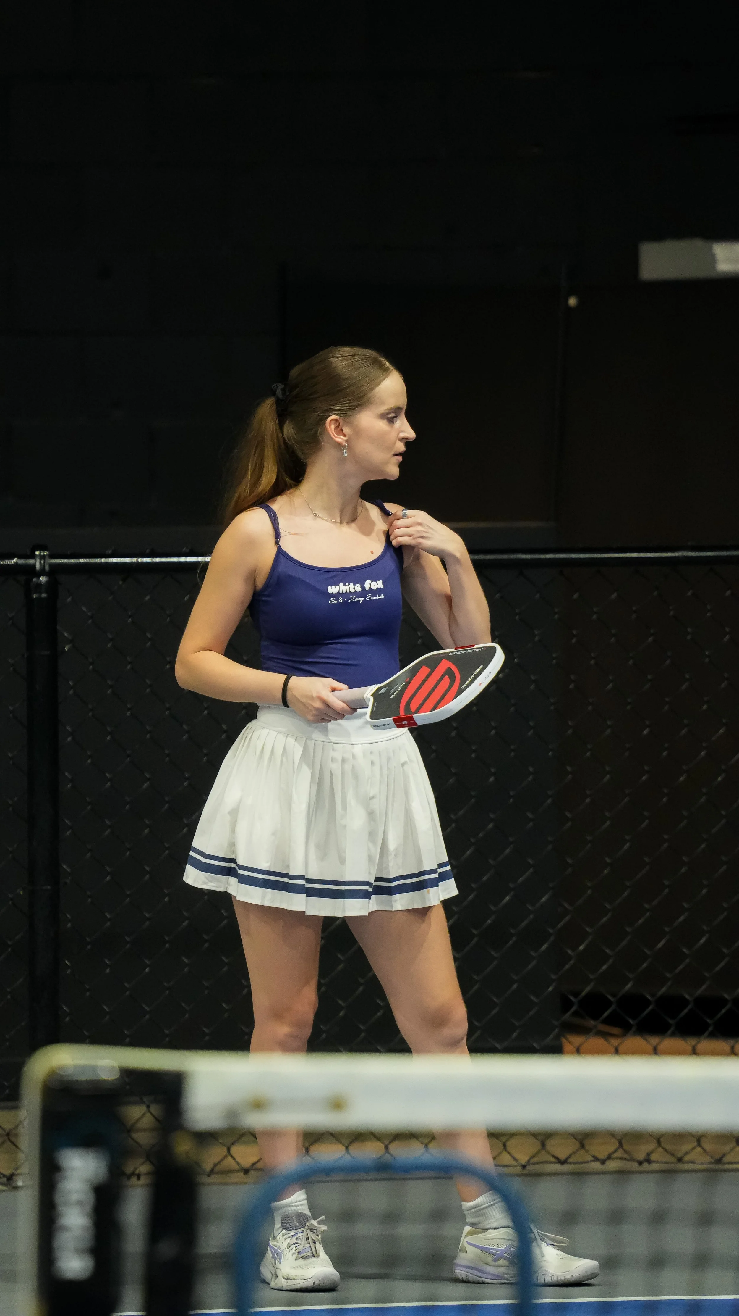 A woman in a blue tank top and white skirt holding a pickleball racket stands near a black fence in an indoor pickleball court.