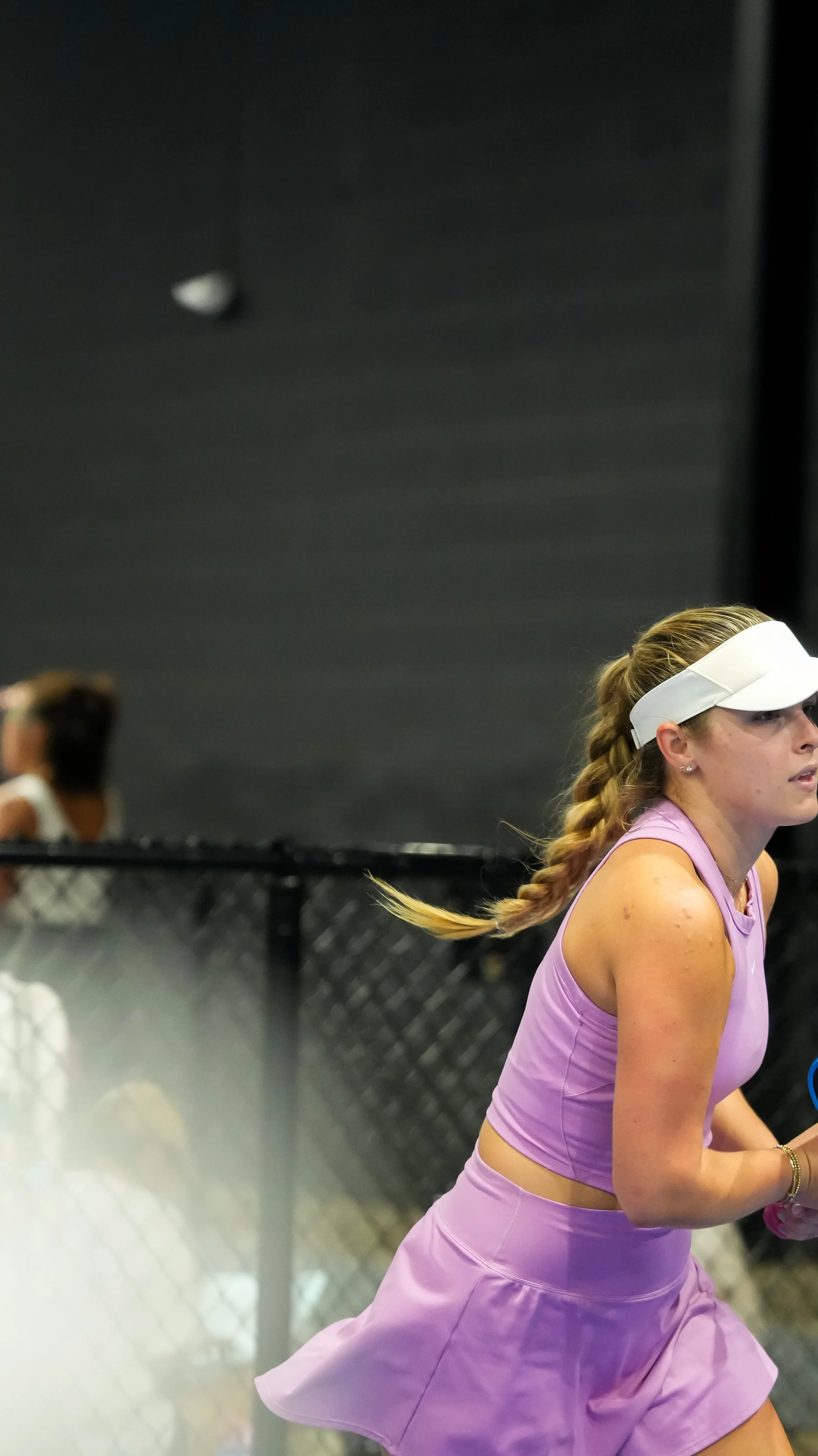 A woman dressed in a pink athletic outfit with a white visor and a braid, appears to be participating in a fitness class or workout session. There are other women in similar outfits in the background, and a black chain-link fence is visible.