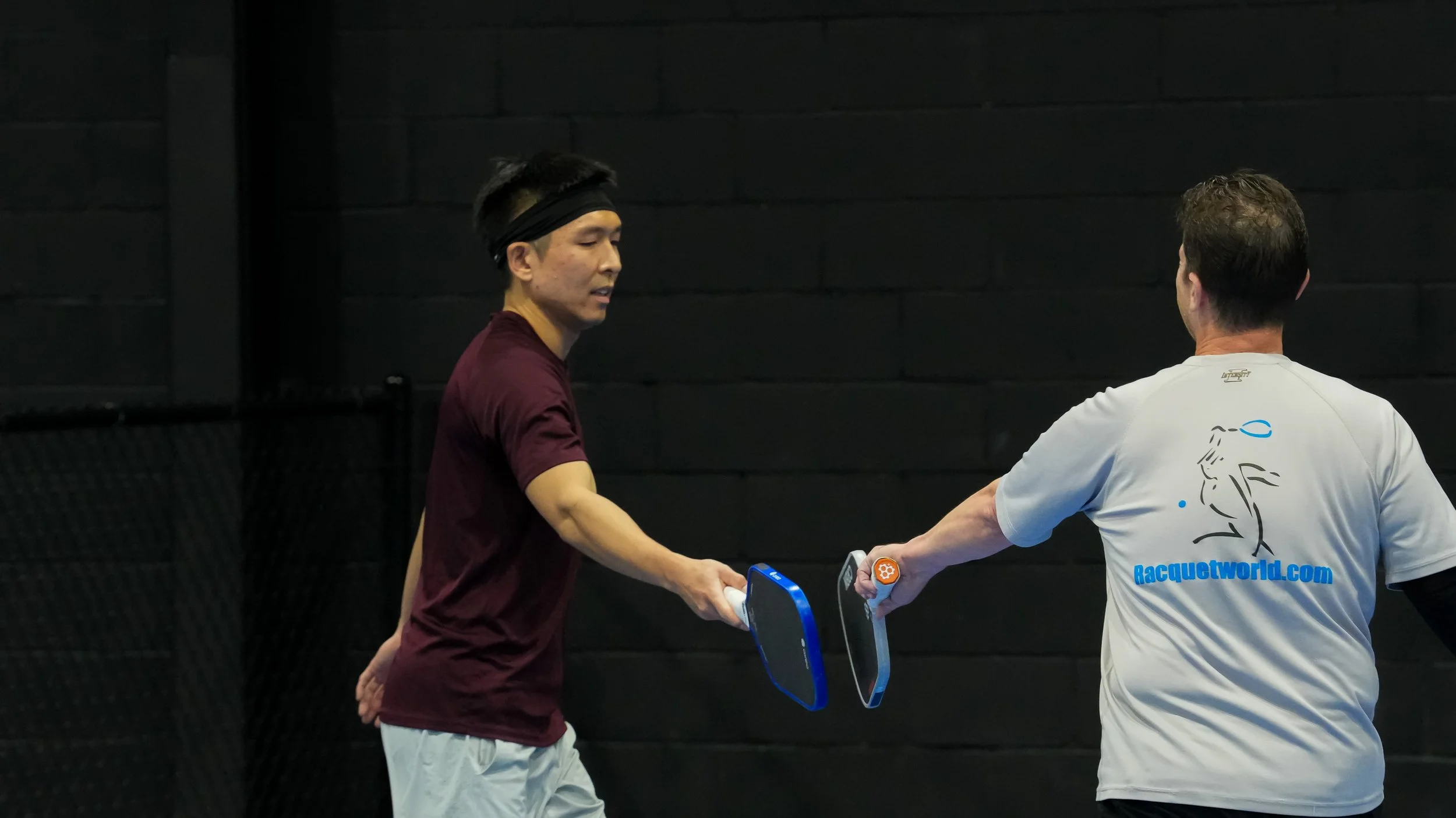 Two men shaking hands in a tennis training facility with a black brick wall background. One man is wearing a maroon shirt and black headband, the other a white tennis shirt with a cartoon face and blue text.