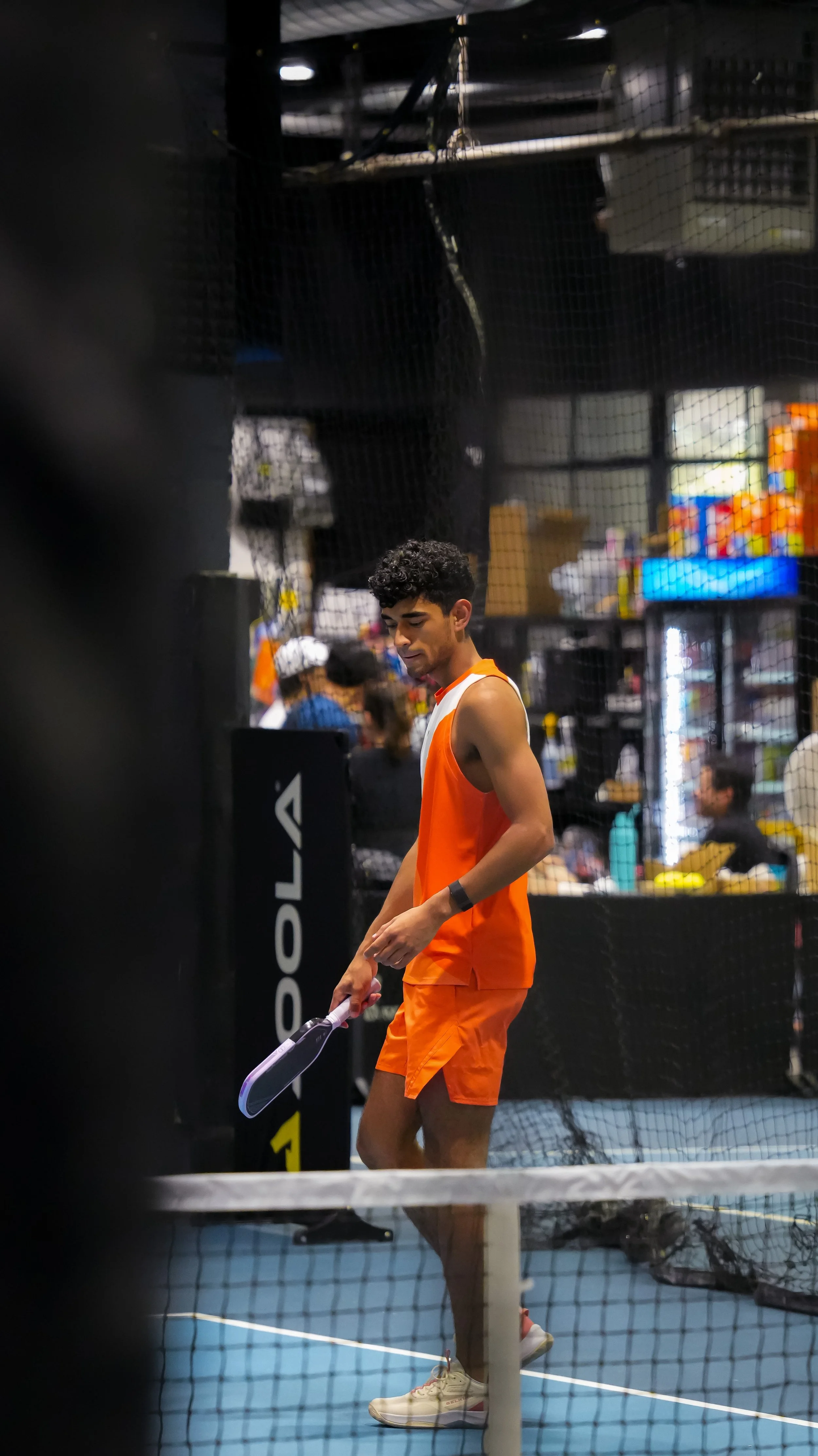 A young man dressed in an orange sports outfit holding a floorball stick on an indoor sports court, with a net in front and explosion of colors in the background.