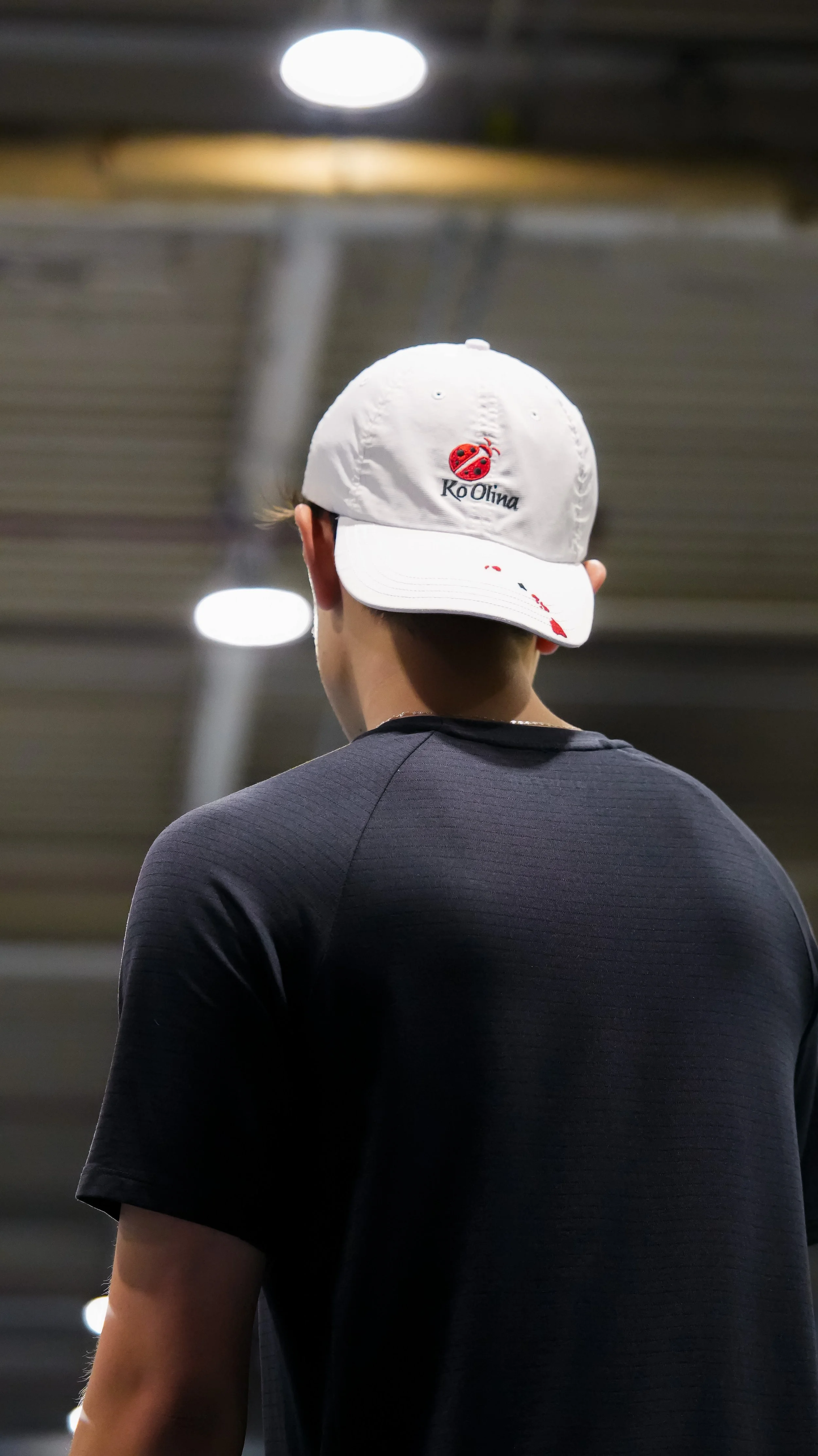 Back of a person wearing a white cap with a small ladybug logo and the text "KoOlina" on it, and a black athletic shirt, in an indoor setting with bright overhead lights.